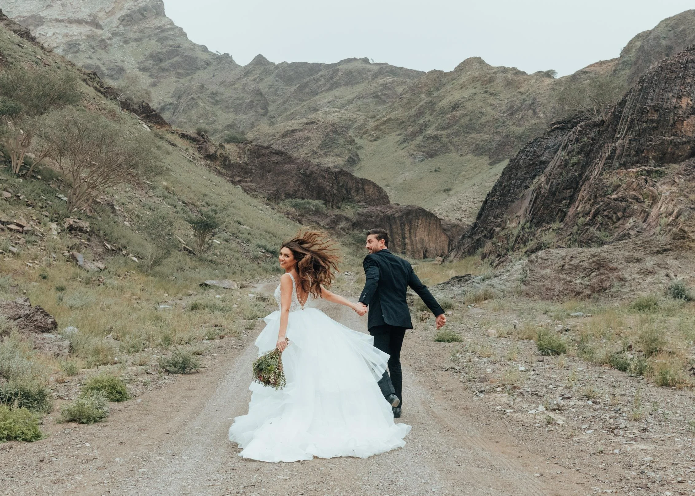 A bride and groom holding hands and dancing on a dirt path in a mountainous landscape. The bride is wearing a white wedding dress and holding a bouquet, while the groom is dressed in a dark suit. They are looking back and smiling.