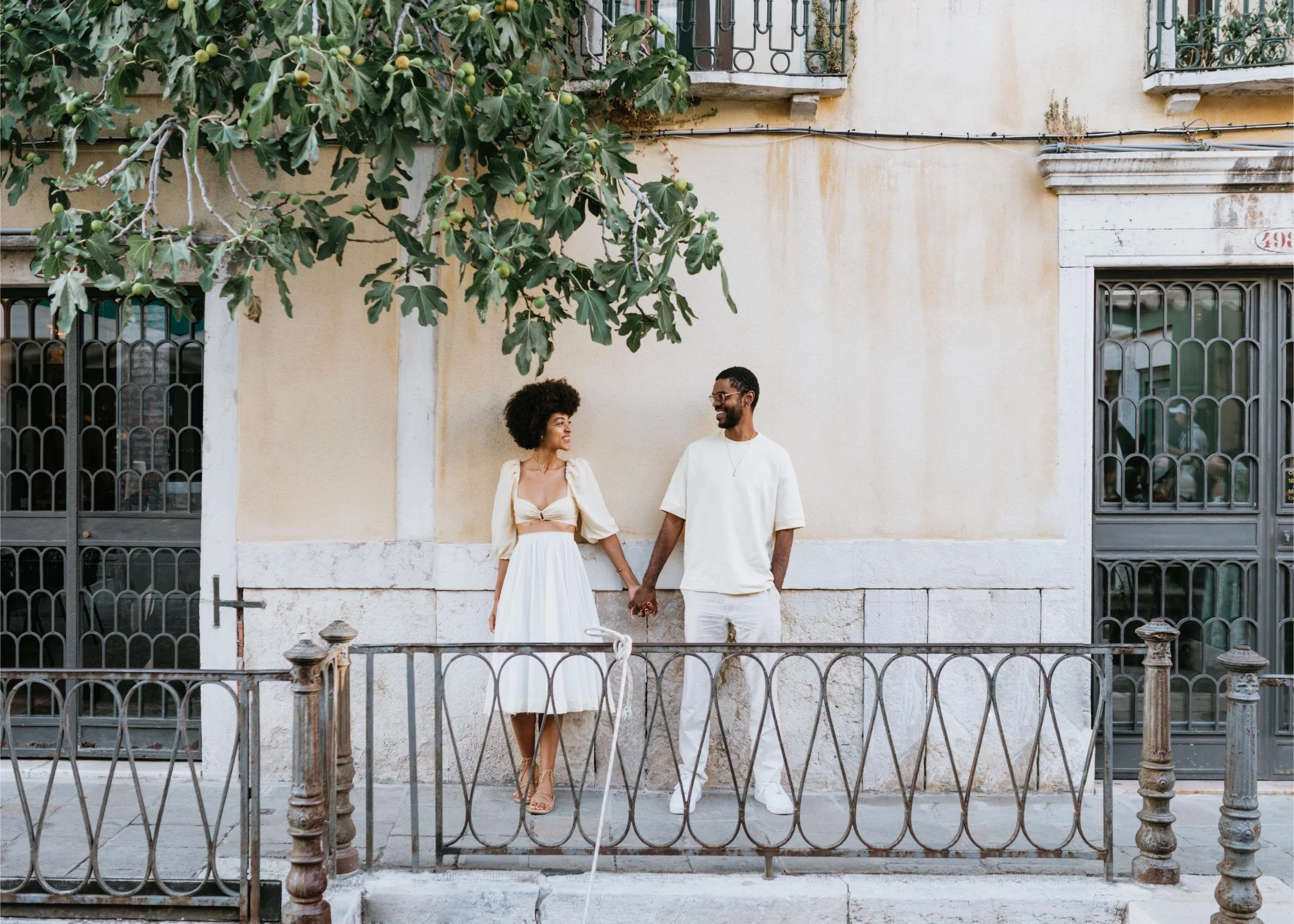 A couple holding hands and smiling at each other standing in front of a beige wall with metal railings and a large leafy tree overhead.