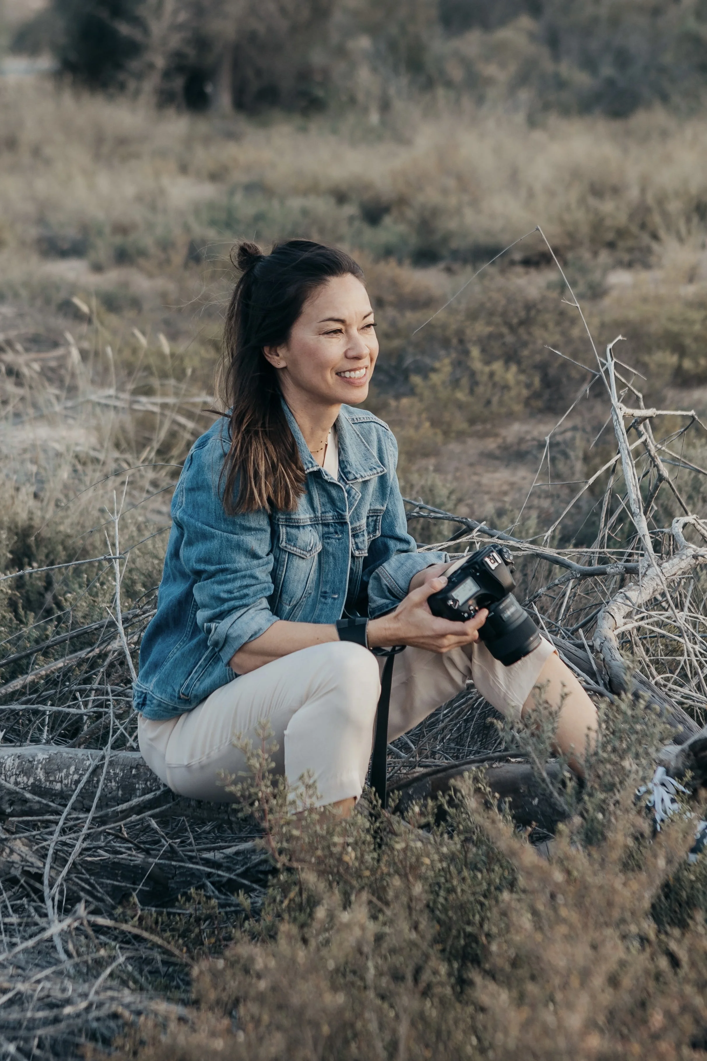 A woman with dark hair tied back, wearing a denim jacket and light-colored pants, sitting amid dry brush and branches outdoors, holding a camera and smiling.