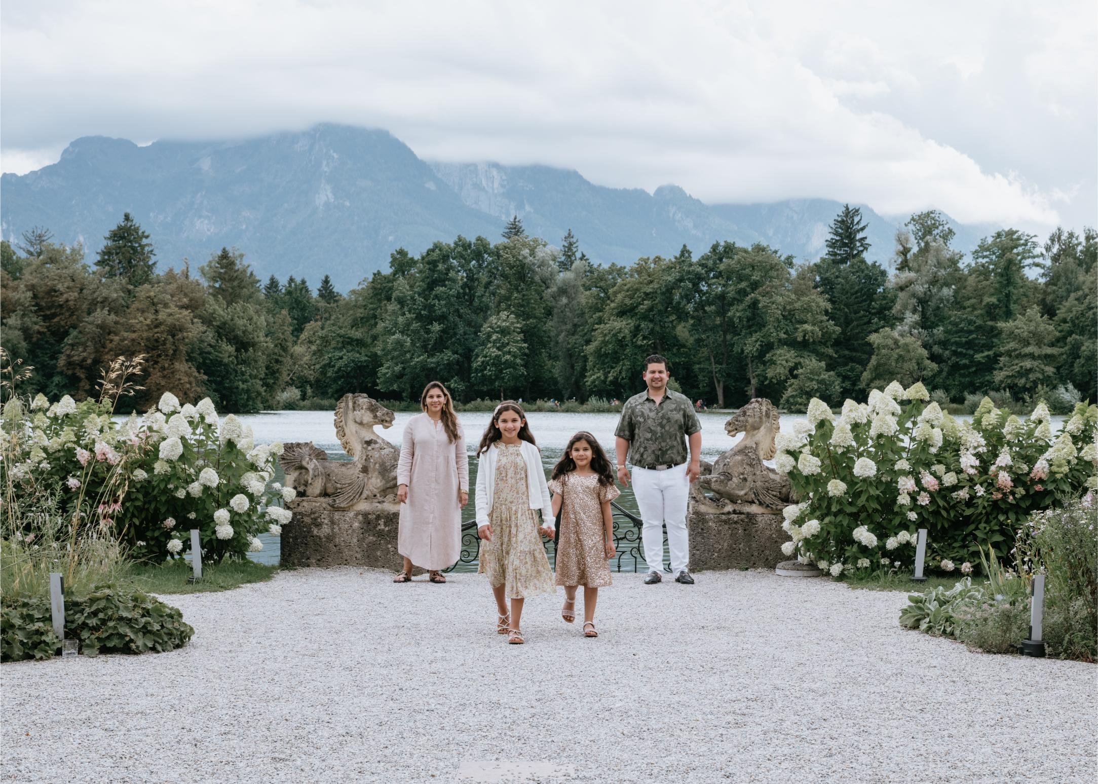 A family of four walking on a gravel path in a garden near a lake with flower bushes and stone lion sculptures, with mountains and cloudy sky in the background.