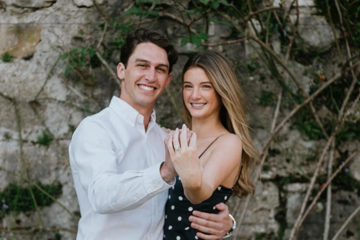 A smiling couple, a man in a white shirt and a woman in a black polka dot dress, holding hands and displaying an engagement ring in front of a stone wall with vines.