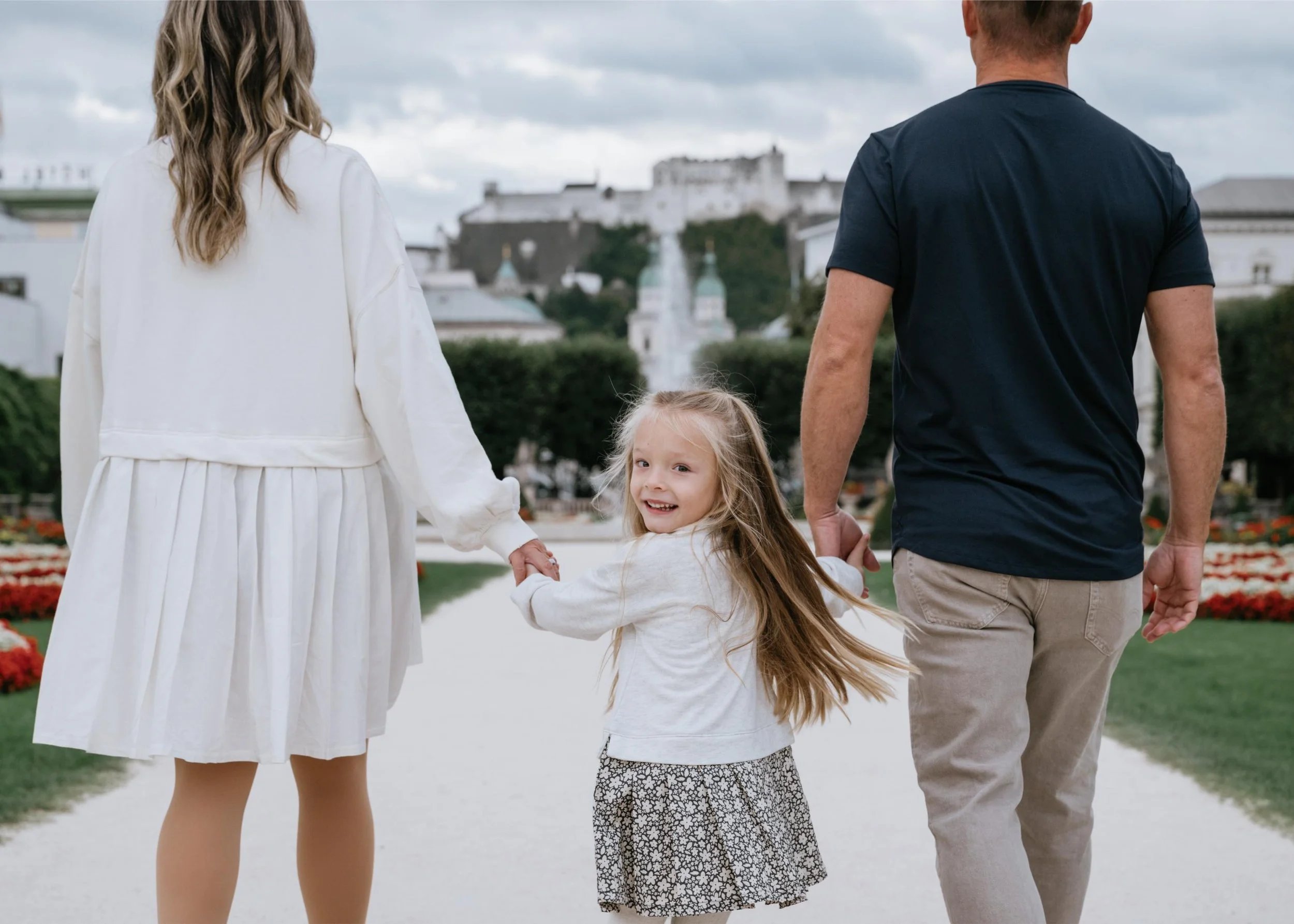A family of three walking hand in hand in a park, with a girl looking back and smiling at the camera, while the parents walk ahead. There are flowers and trees in the background.