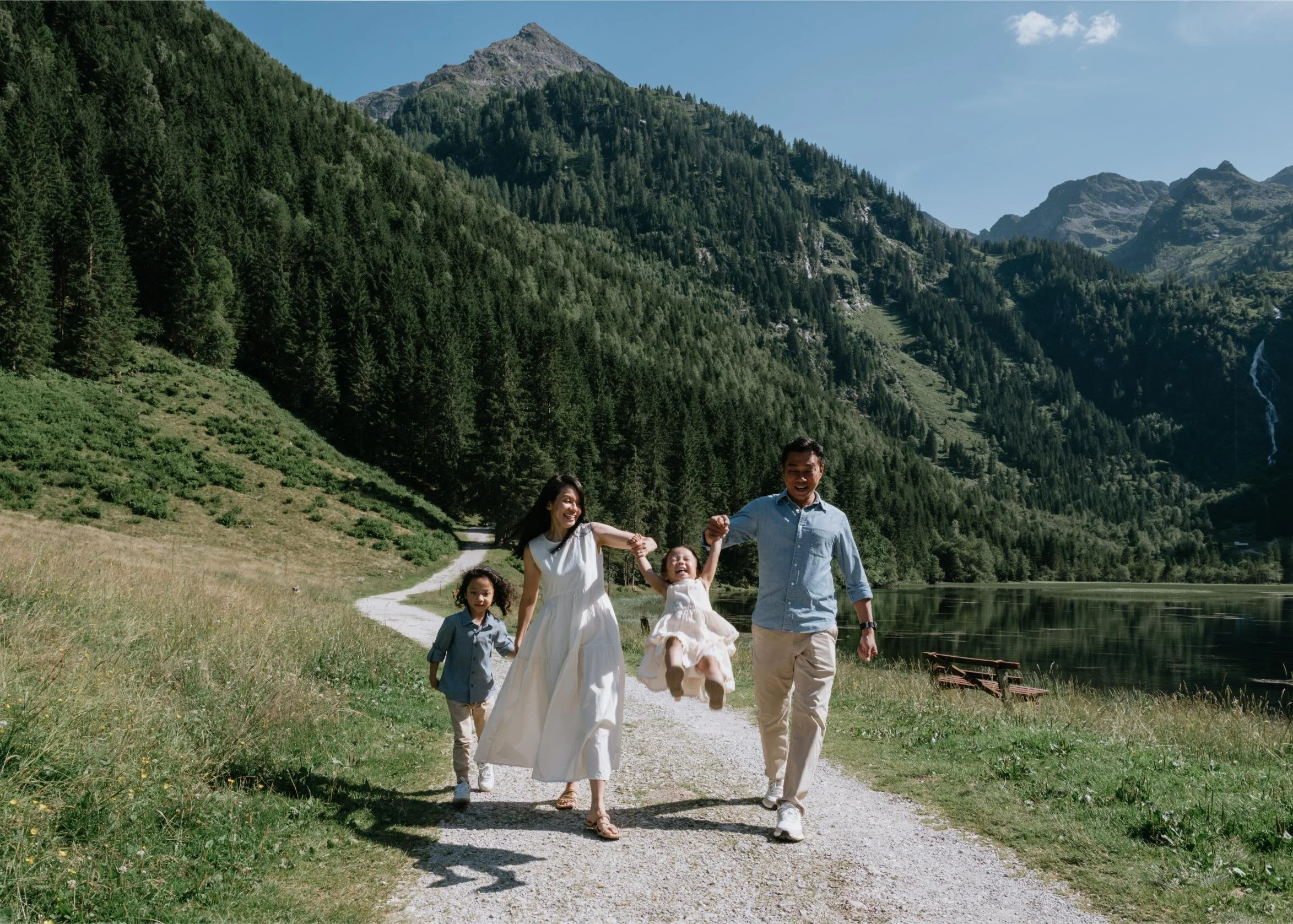 A family of five walking on a gravel path near a lake, surrounded by lush green mountains and trees, with sunny weather