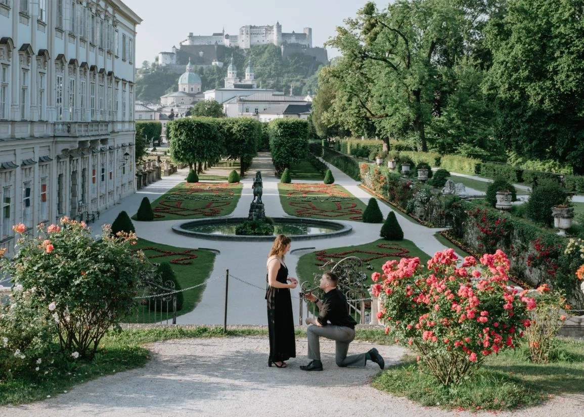 A man is proposing marriage to a woman in a beautiful formal garden with neatly trimmed bushes, a central fountain, and colorful flowers, with a castle on a hill in the background.