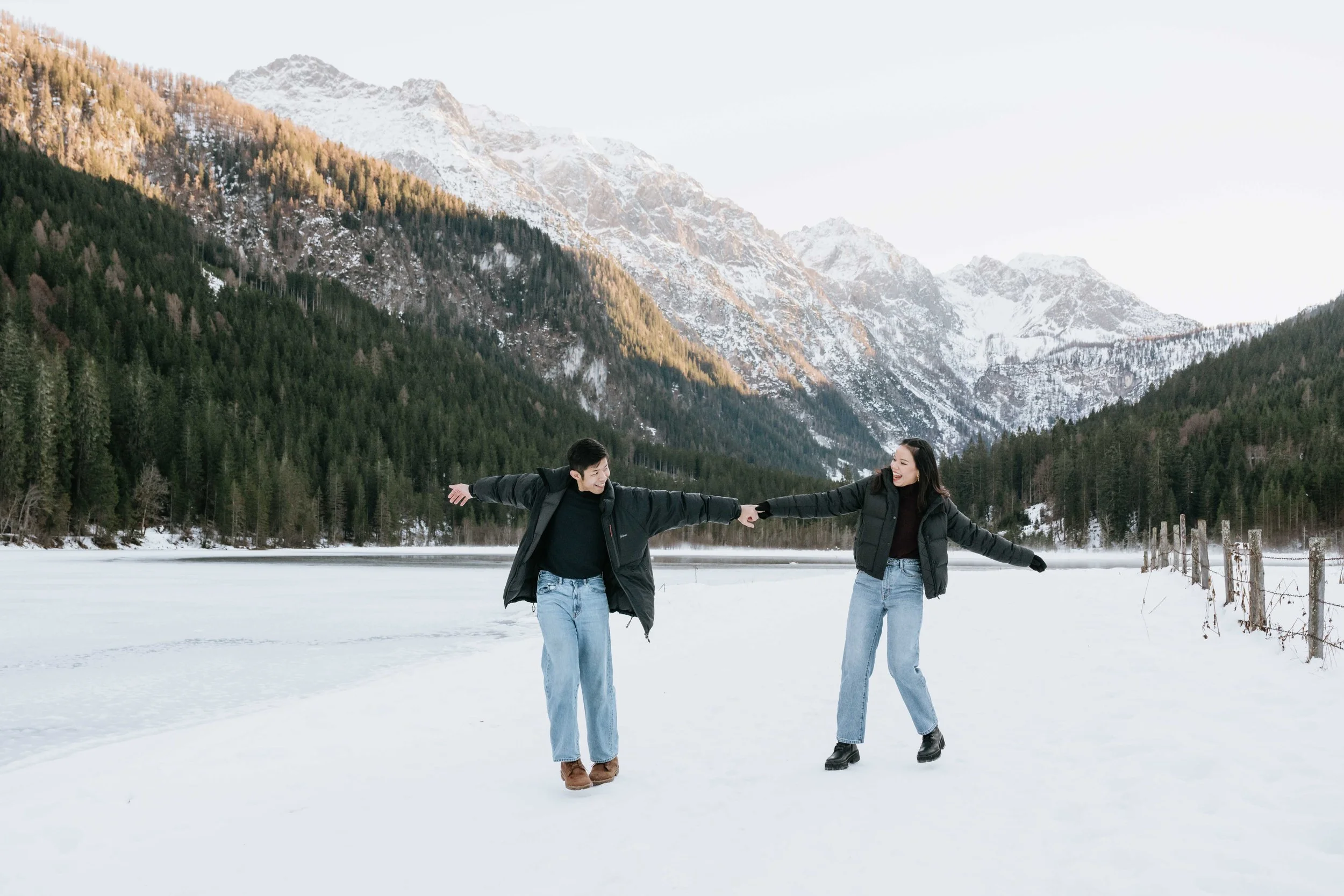 A couple holding hands while dancing on snow-covered ground in front of mountains and pine trees during winter.