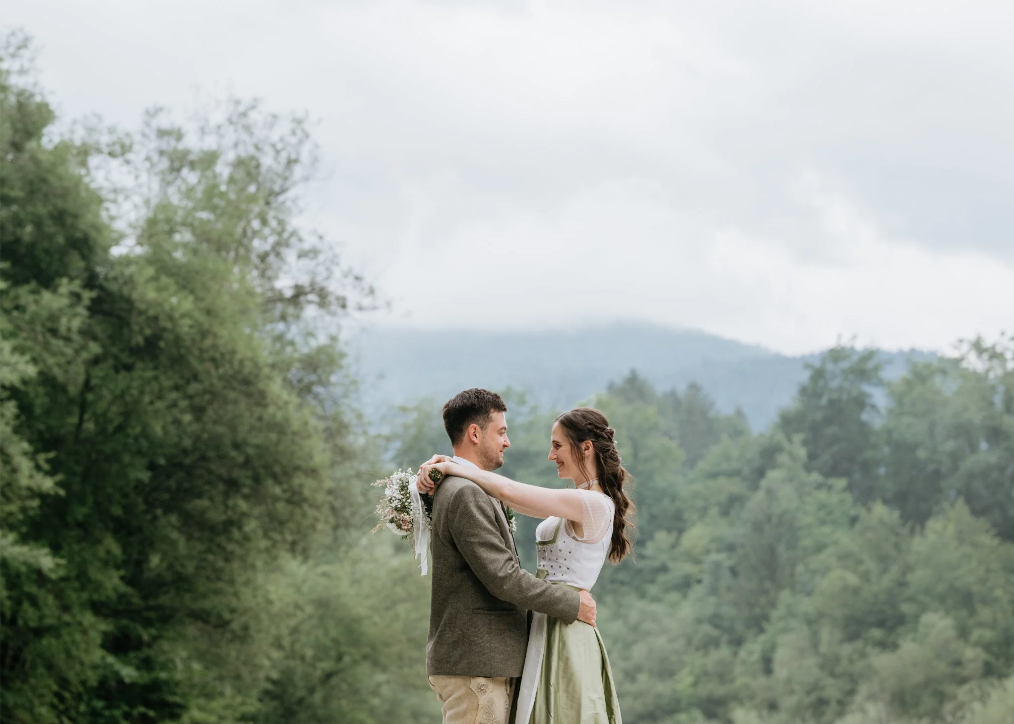A bride and groom embrace outdoors in a green, wooded landscape with mountain range in the background.