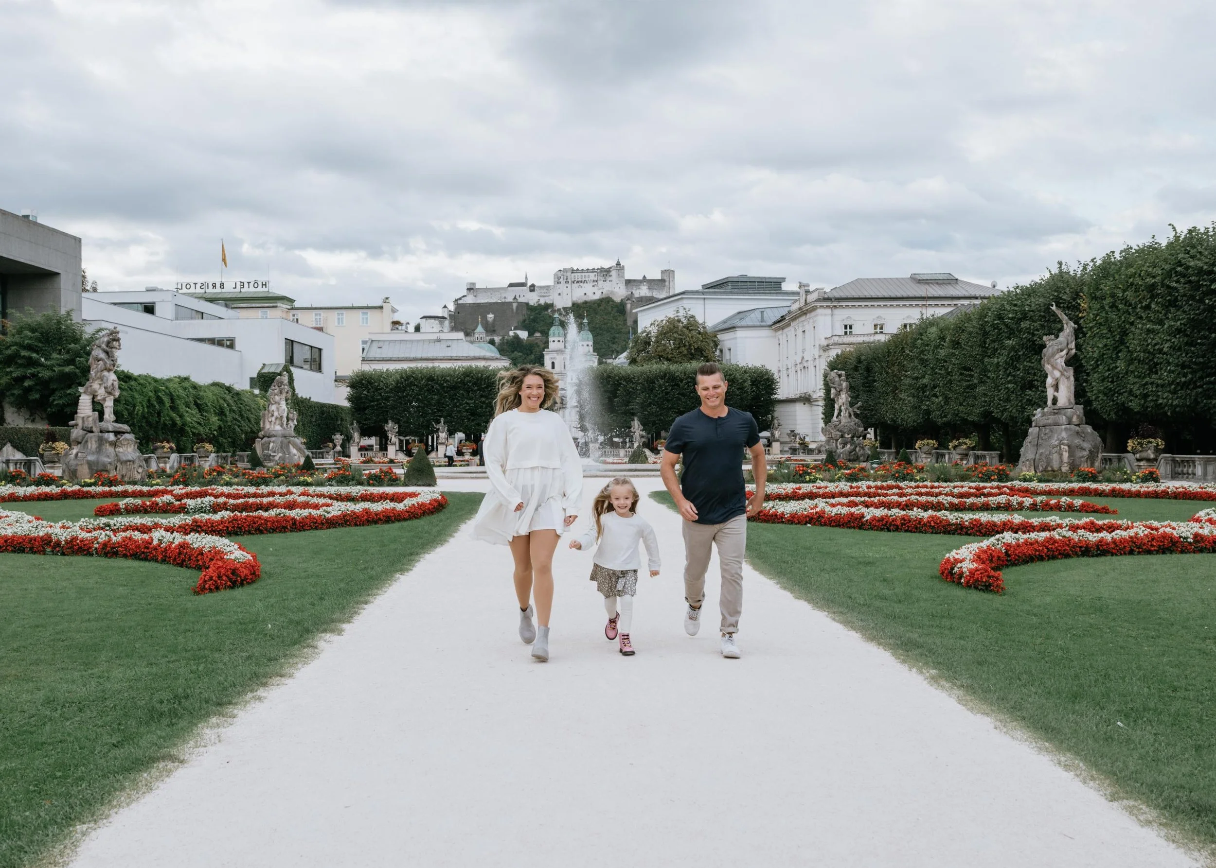 Family of three, two adults and a child, running and smiling on a path in a landscaped park with statues, fountains, and historic buildings in the background.