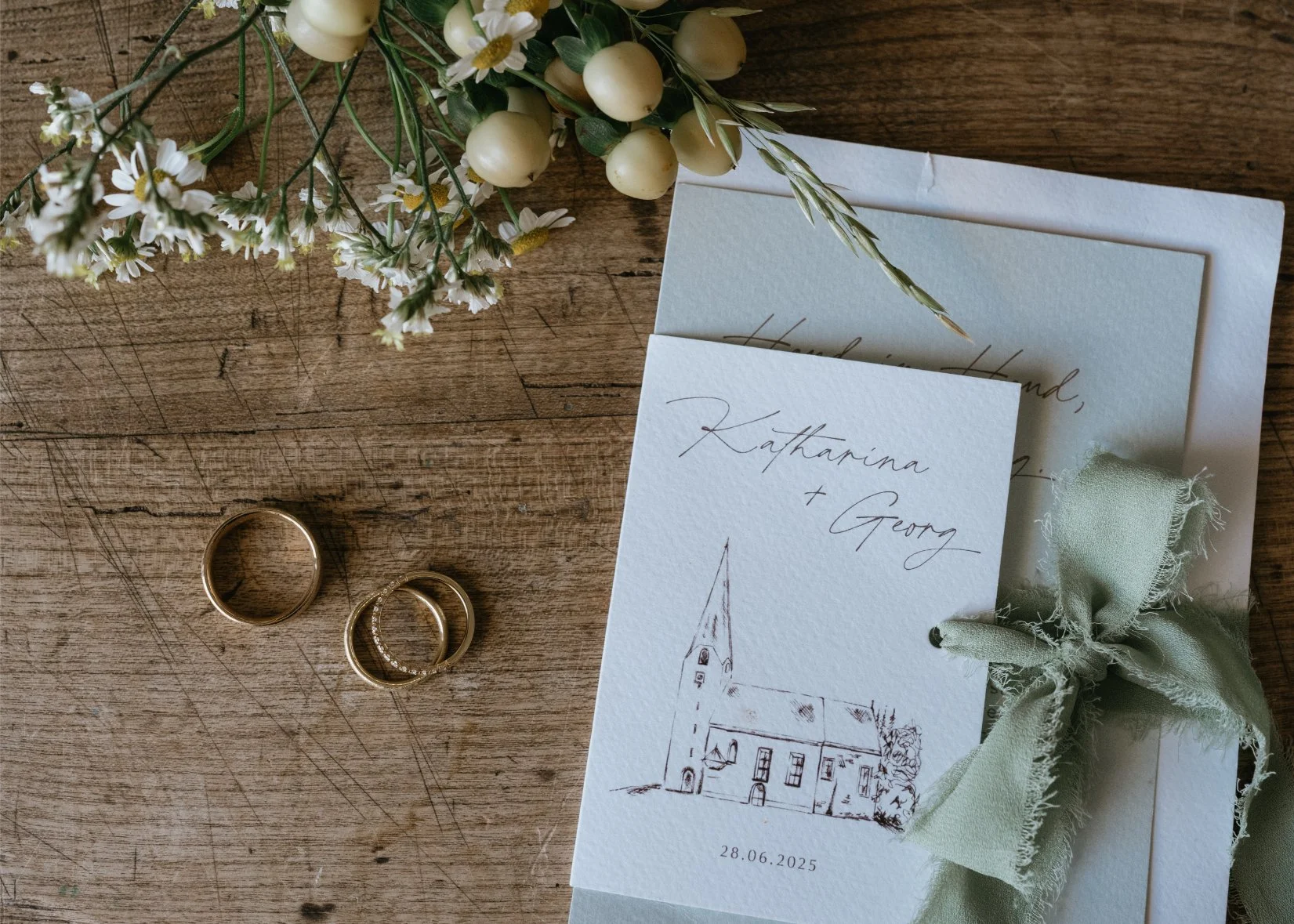 Wedding invitation with sketch of a church, a green bow, and the names Katharina and Georg, date 28.06.2025, arranged on a wooden surface with white flowers and two gold rings nearby.