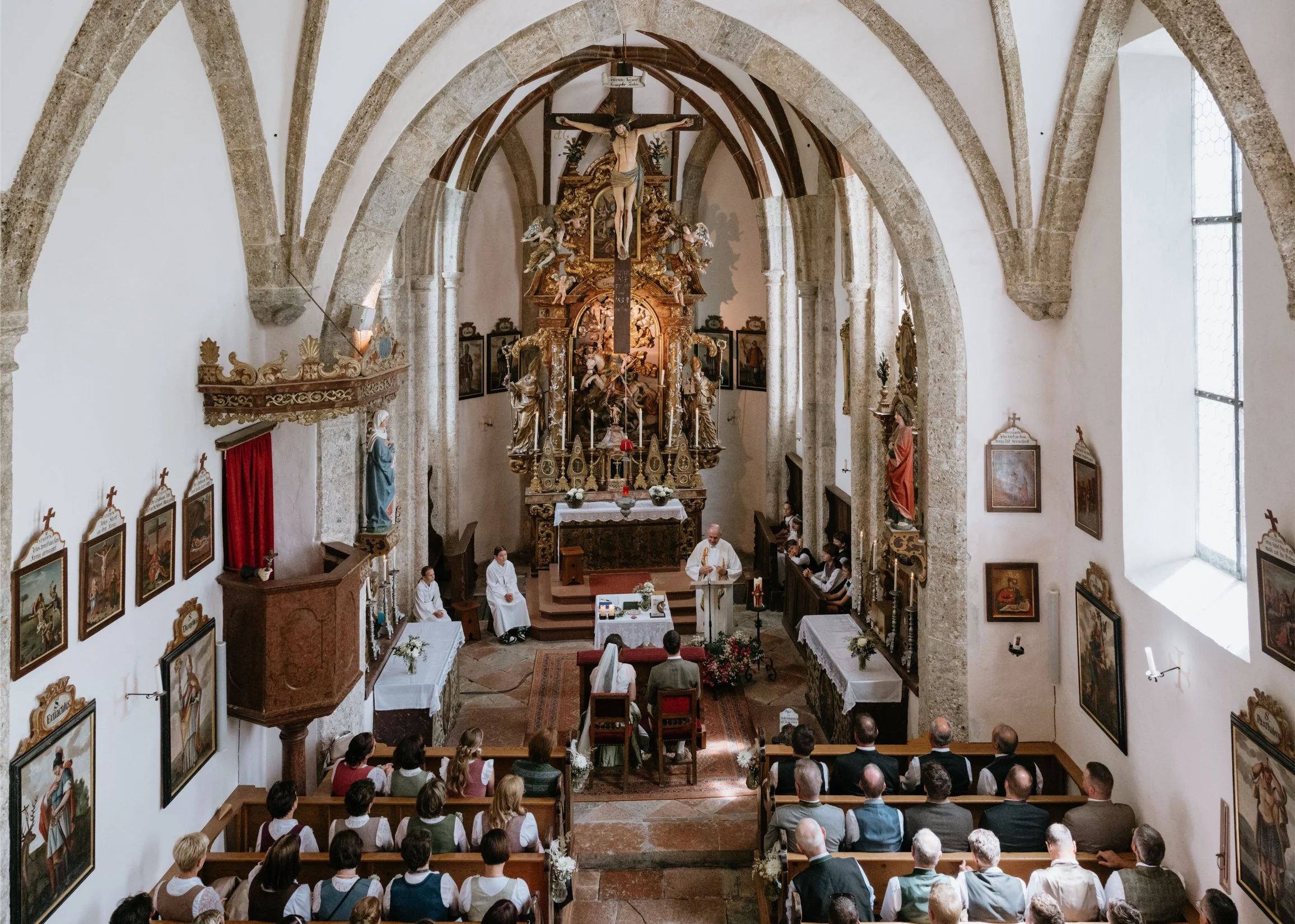 Interior of a church during a wedding ceremony, with the bride and groom seated in front of the altar and people in pews watching. The priest stands at a lectern, and the church has ornate religious artwork and sculptures, tall arched windows, and st