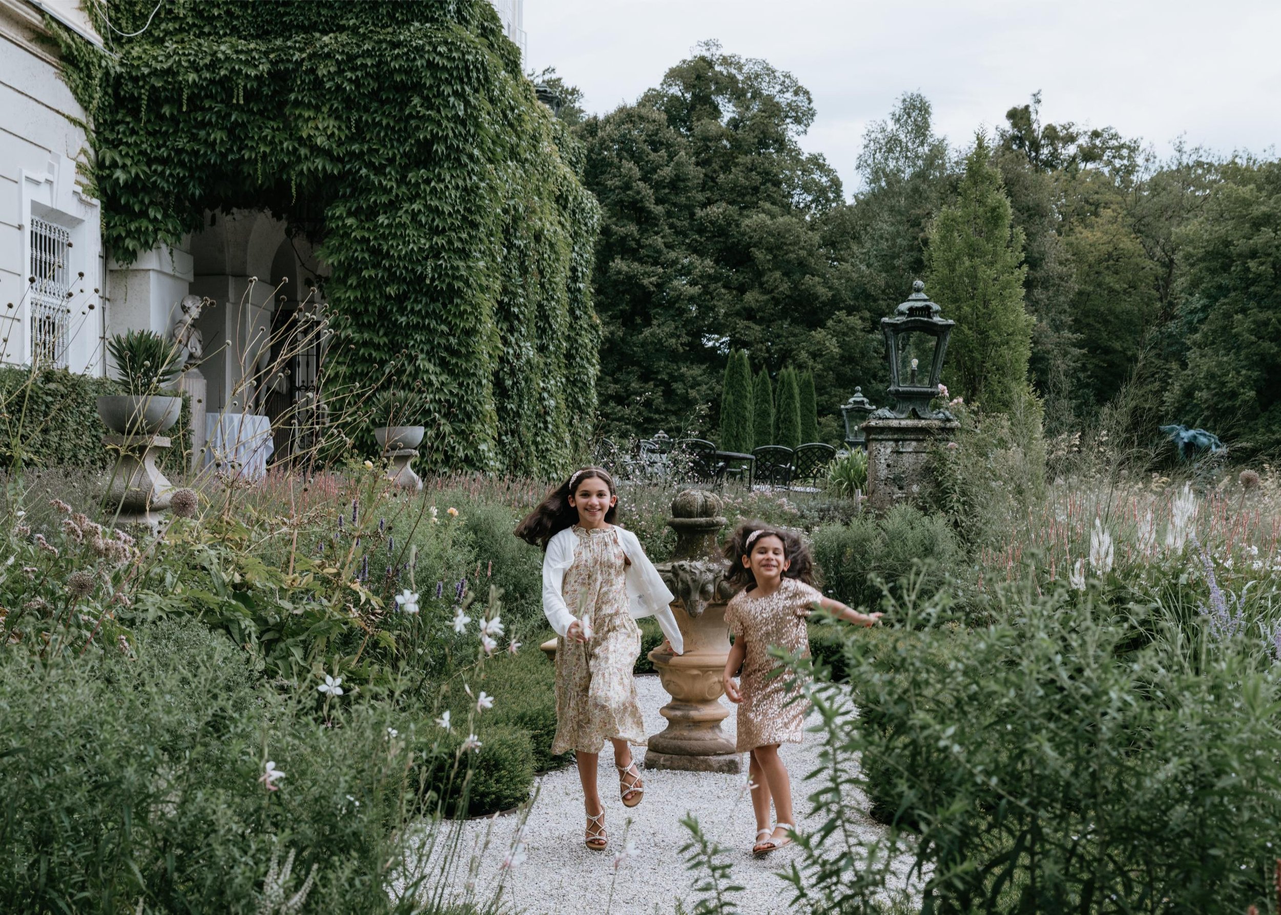 Two young girls running and smiling in a lush garden with various plants, flowers, and decorative garden features. They appear to be enjoying a sunny day.