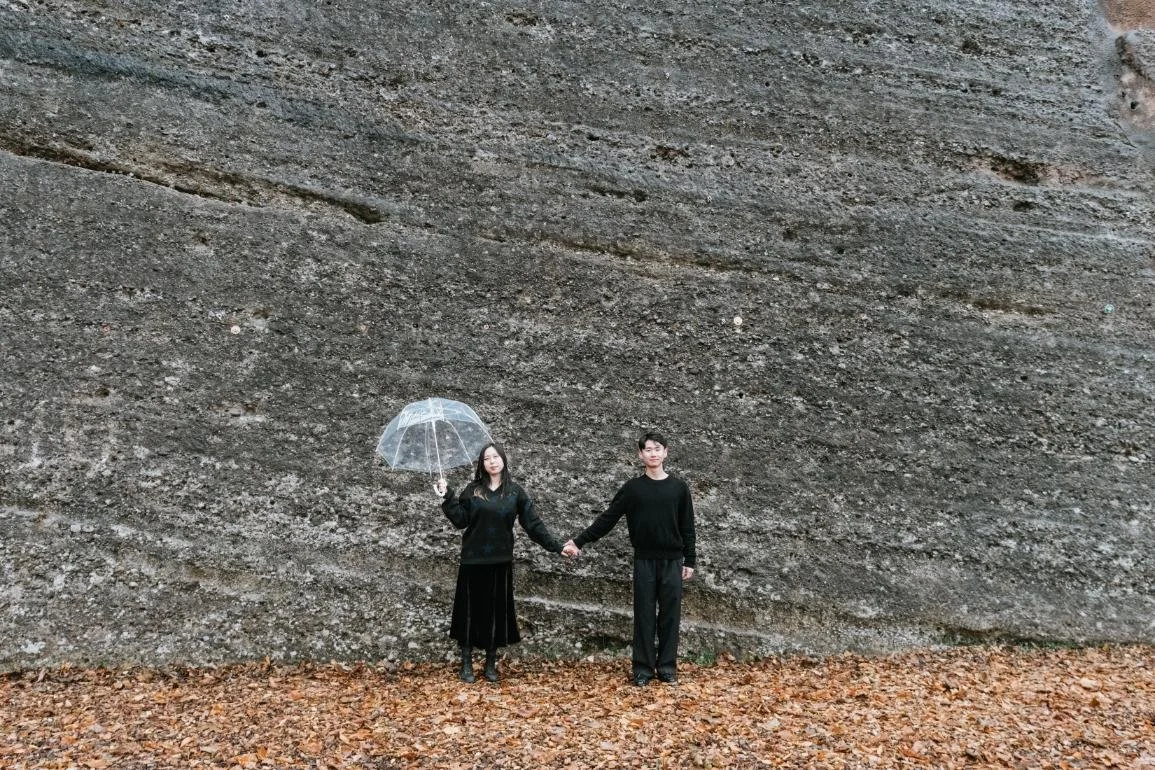 A man and woman holding hands, with the woman holding a transparent umbrella, standing against a large sloped rock wall surrounded by fallen leaves.