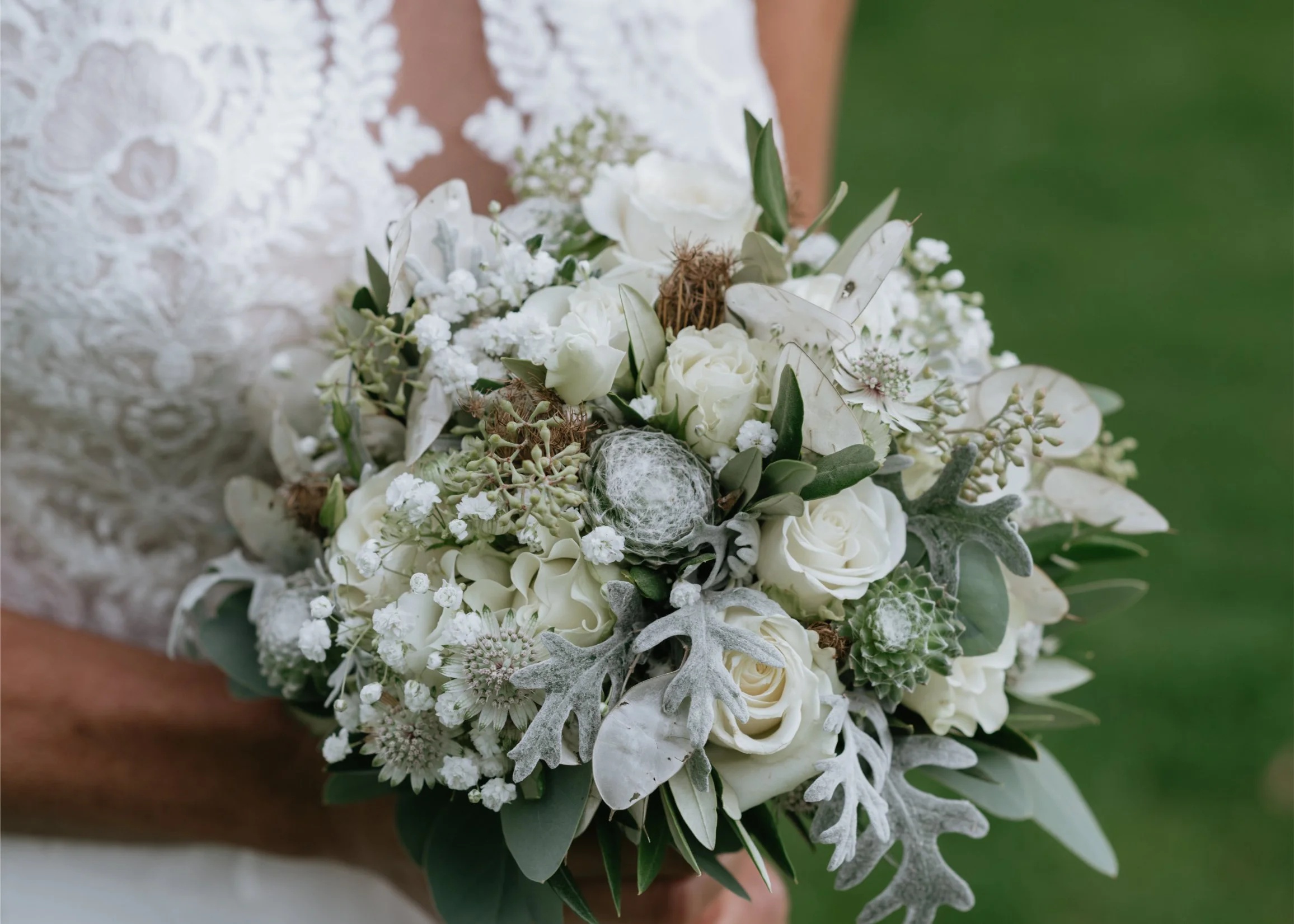 A bride holding a white floral wedding bouquet with roses, greenery, and decorative accents like dried seed pods and silk spider web details in an outdoor setting.
