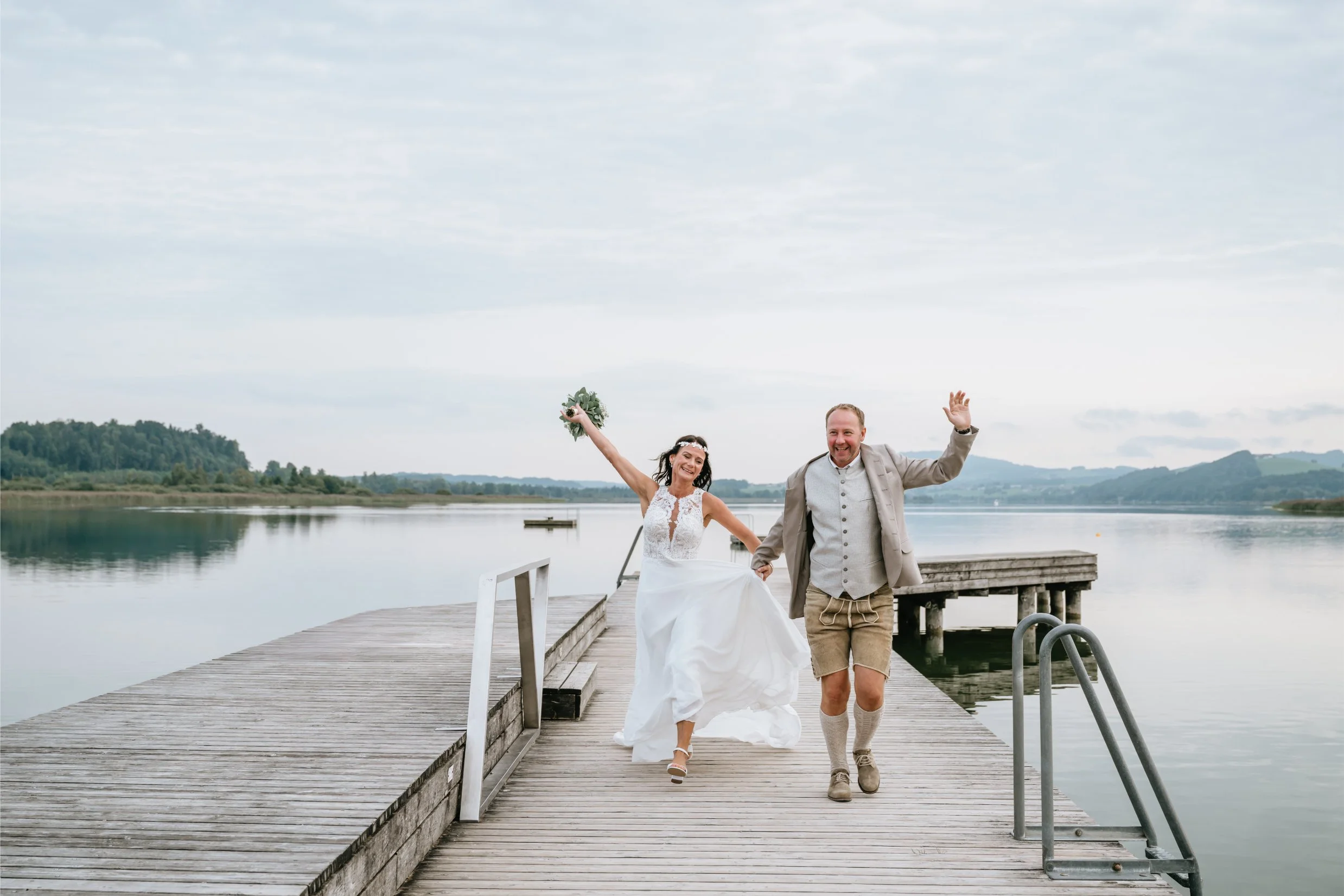 A smiling bride and groom running hand in hand on a wooden dock by a calm lake, with a cloudy sky above and distant hills in the background, celebrating their wedding.