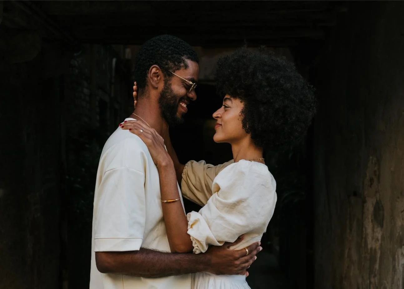 A happy couple, a man with glasses and a woman with natural curly hair, looking into each other's eyes and smiling, in an intimate embrace.