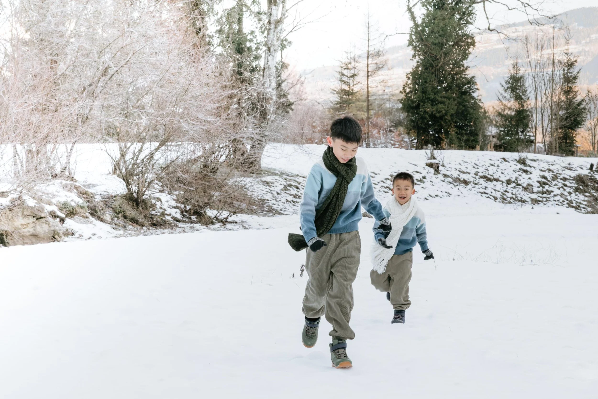 Two young boys in winter clothes playing and running on snow-covered ground outdoors, with trees and mountains in the background.