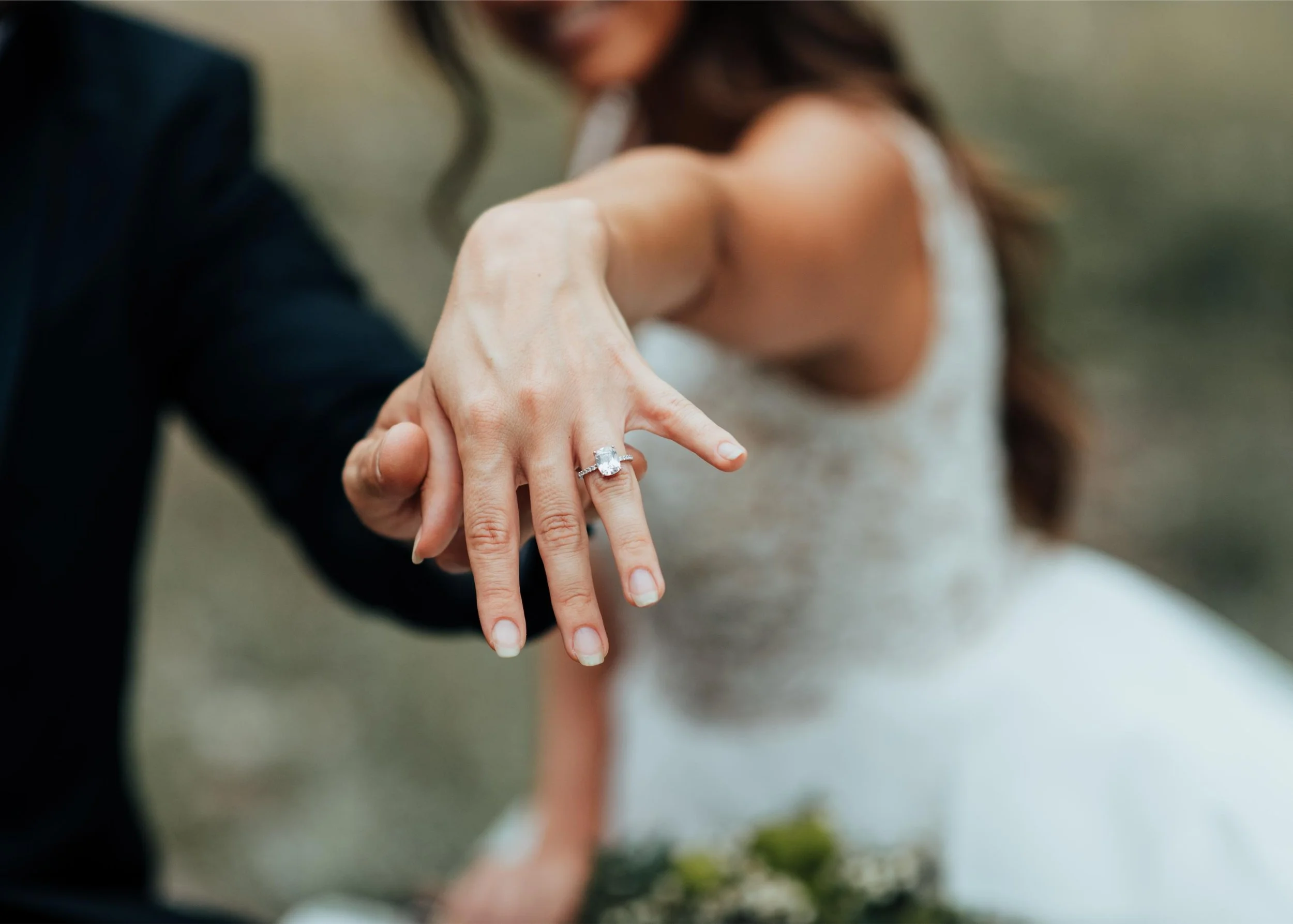 Close-up of a woman's hand showing an engagement ring with a large center diamond. A person in a dark suit is holding her hand. The background shows a blurred woman in a wedding dress, likely a bride.
