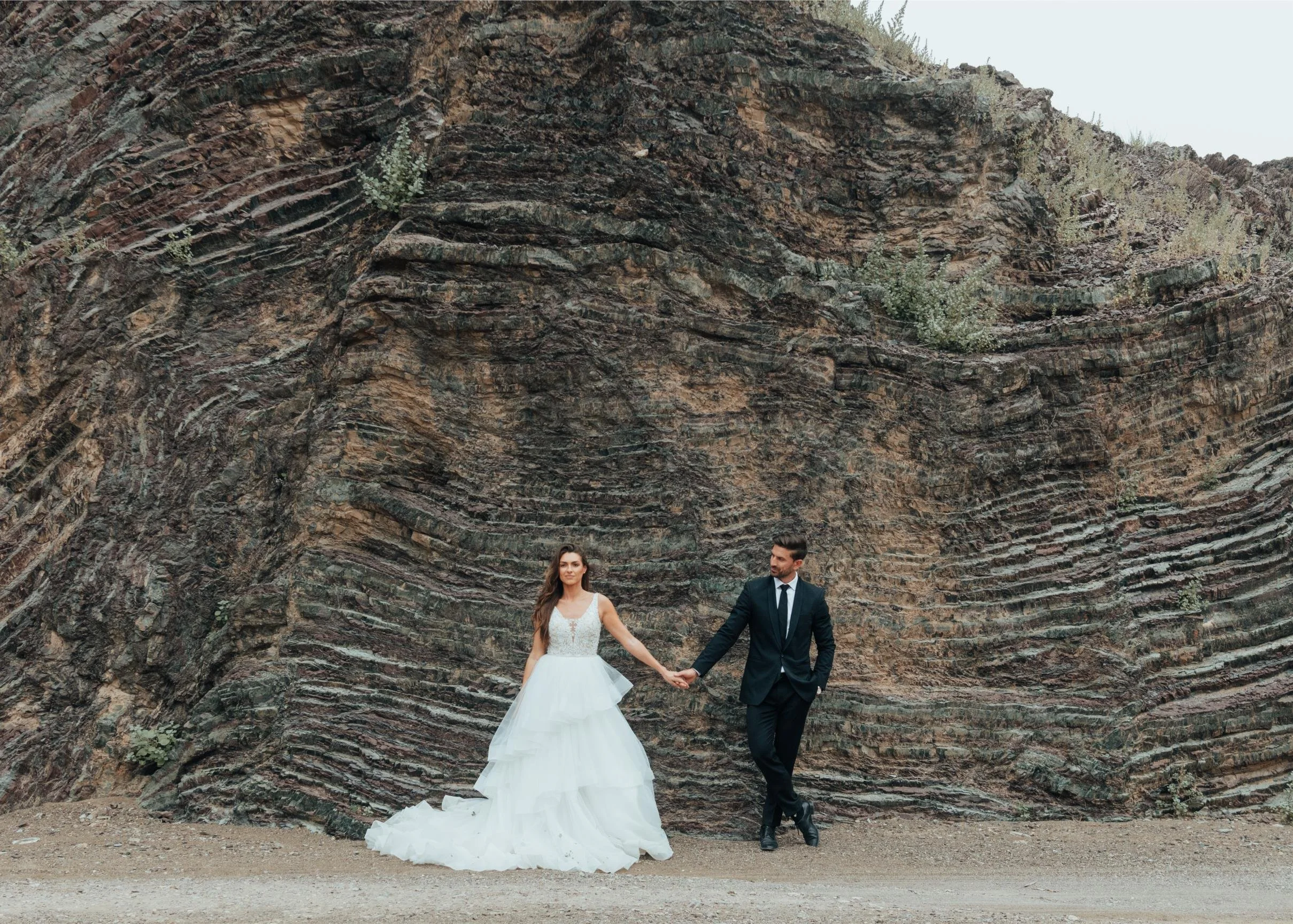A bride in a white wedding gown and a groom in a black suit holding hands and dancing in front of a large, layered rock formation.