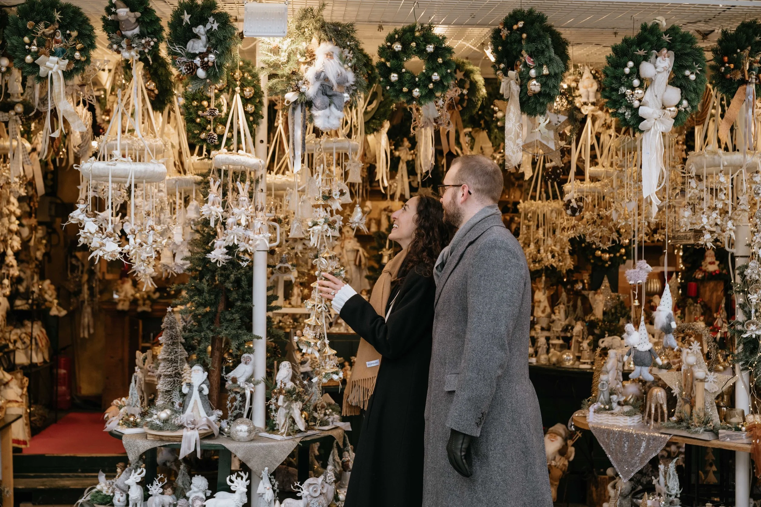 Two people, a woman and a man, shopping for Christmas decorations at an ornament store with a wide variety of holiday ornaments, wreaths, and festive decor in the background.