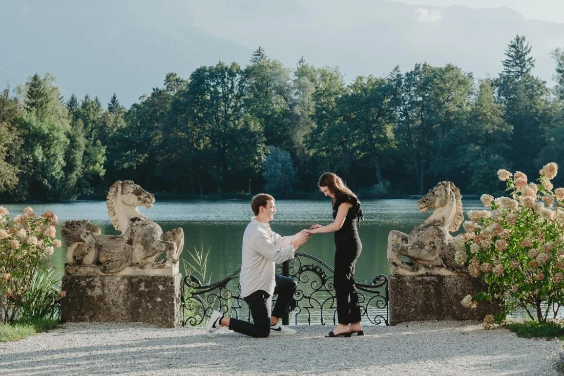 A man proposes to a woman by a lake, surrounded by greenery and stone sculptures of horses.