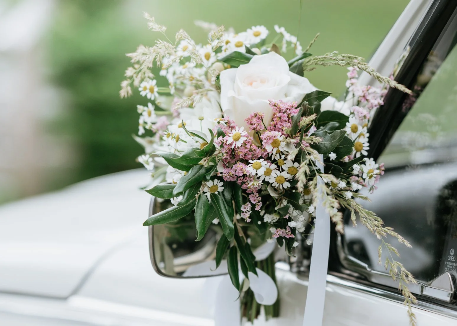 A bouquet of white and pink flowers attached to the side mirror of a white vehicle, with a blurred green background.