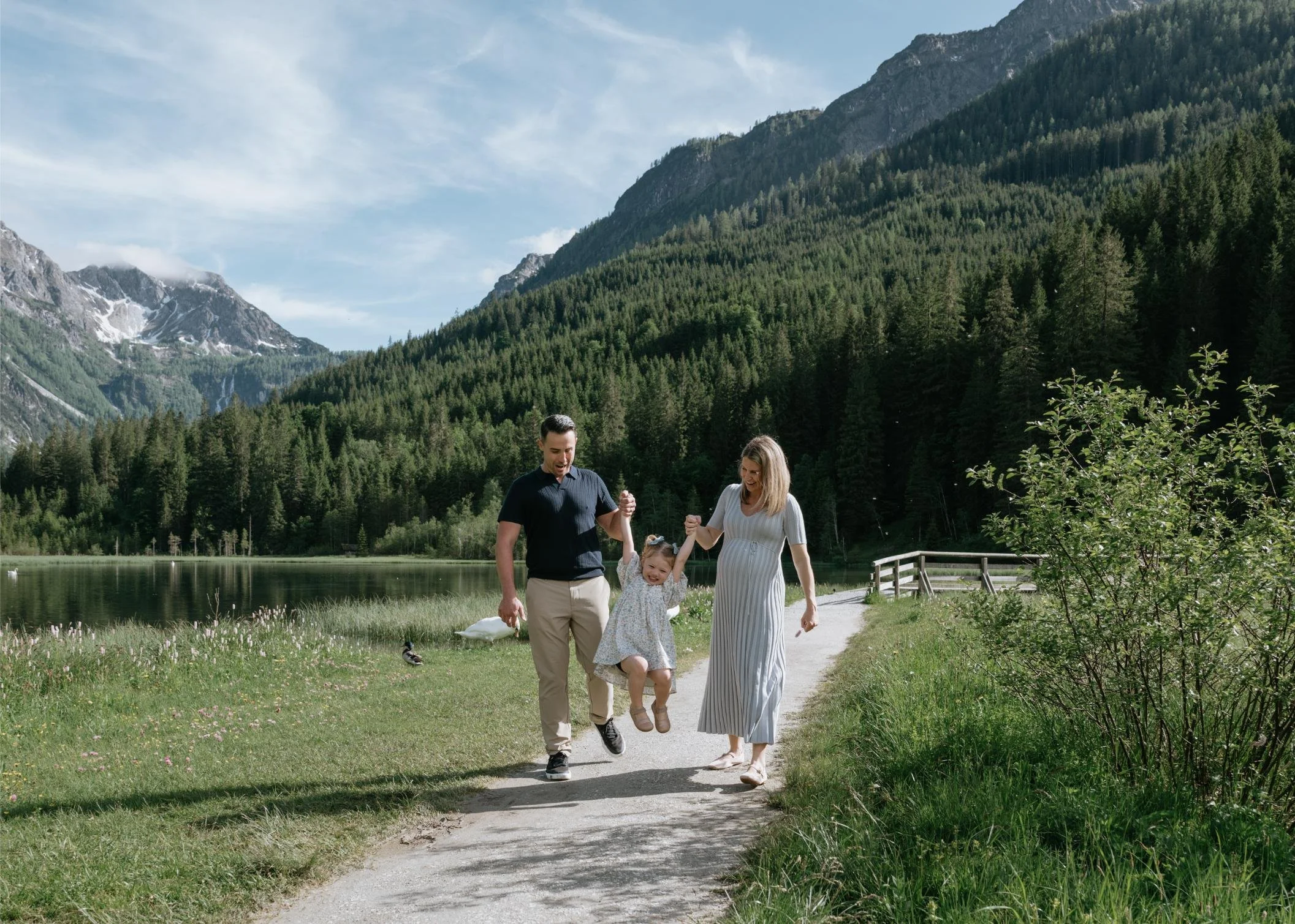 A family of three enjoying a walk outdoors near a lake and mountains, with the child jumping in the air and the parents holding hands.