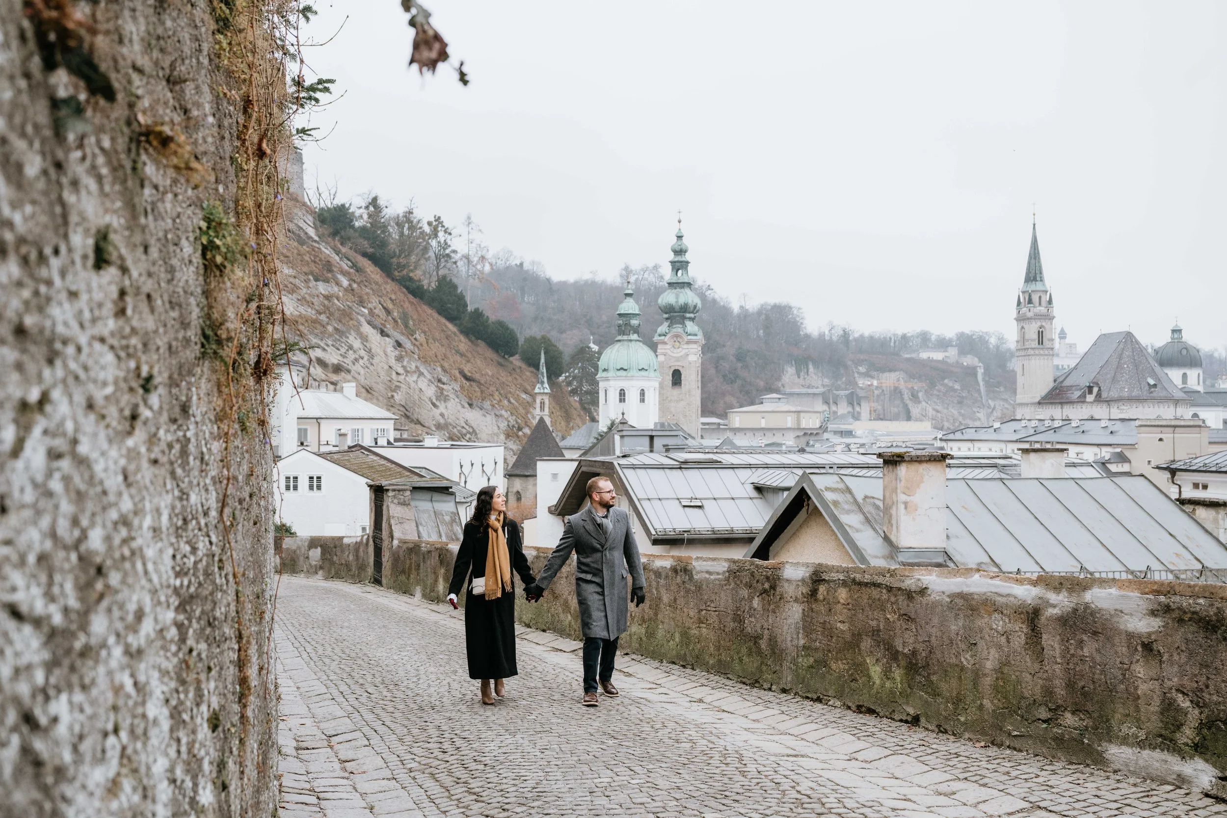 A couple walking hand in hand on a cobblestone street with European-style buildings and church steeples in the background.