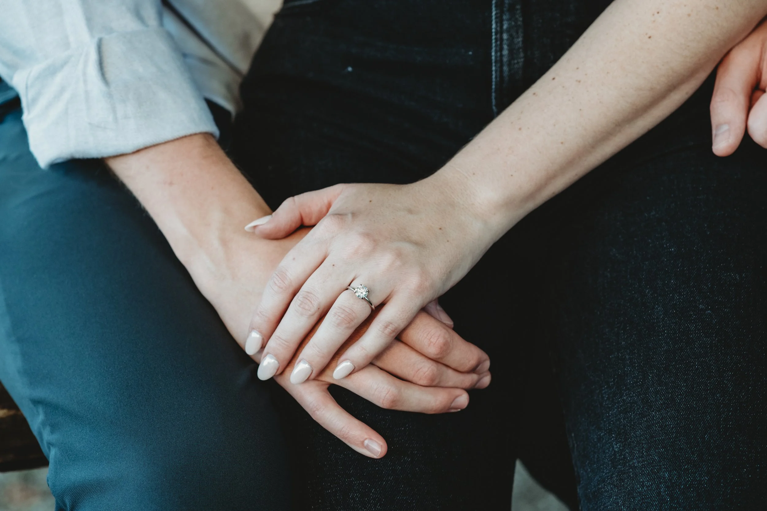 Close-up of a woman's hand with an engagement ring resting on a man's hand, both sitting close together.