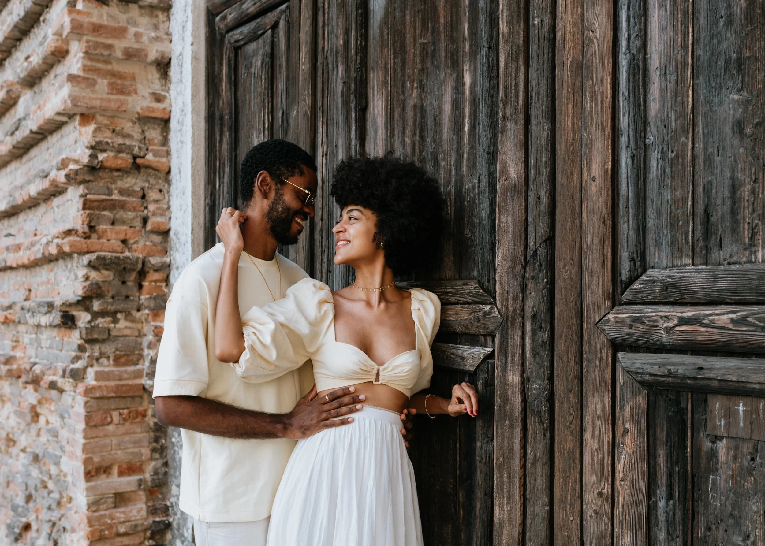 A couple standing close to each other, smiling and looking into each other's eyes, in front of a rustic wooden door.