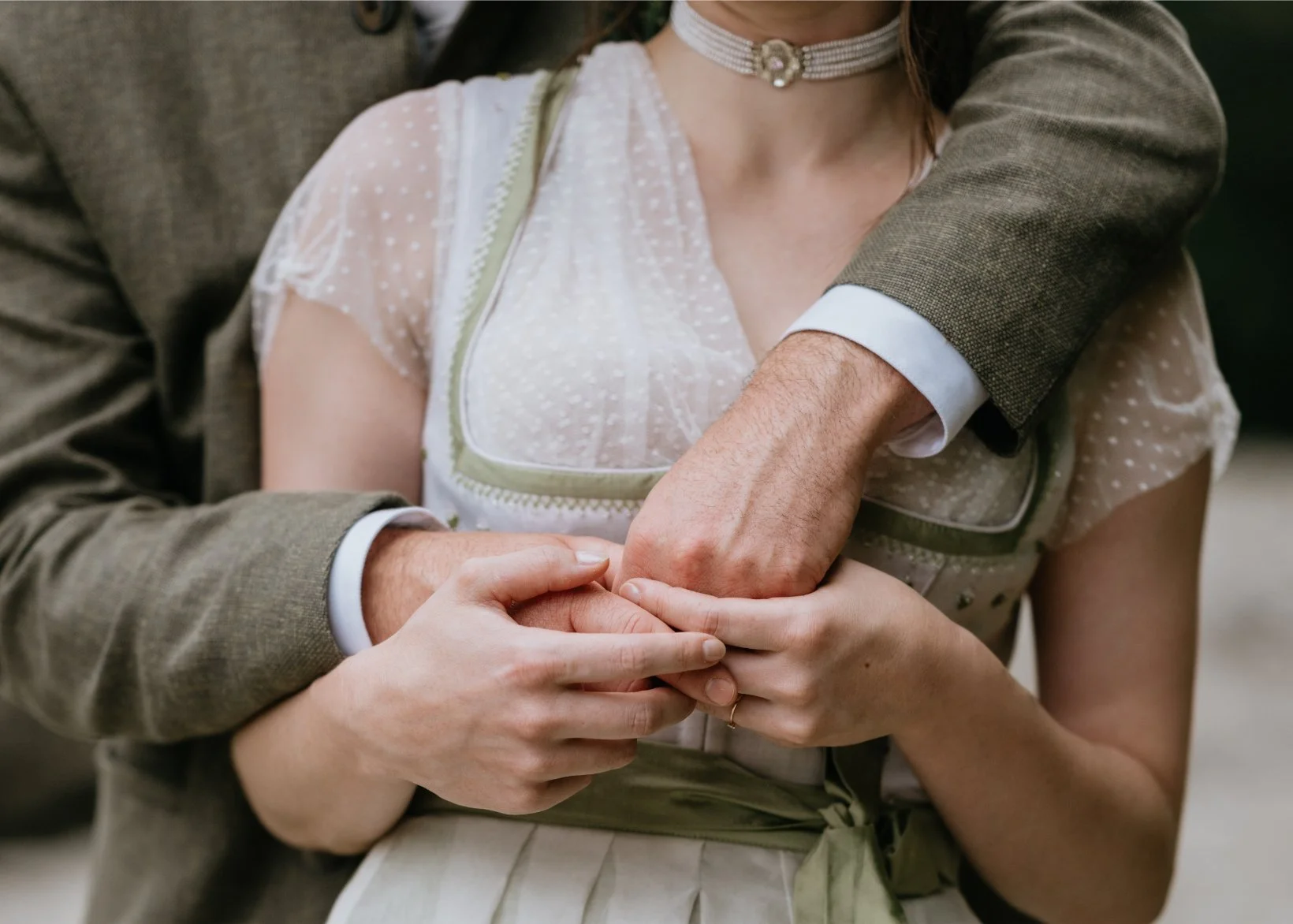 A couple holding hands, with the woman's hand on top of the man's hand. The woman is wearing a vintage-style dress with sheer polka dot fabric and a choker necklace. The man's arm is wrapped around the woman, wearing a brown suit jacket.