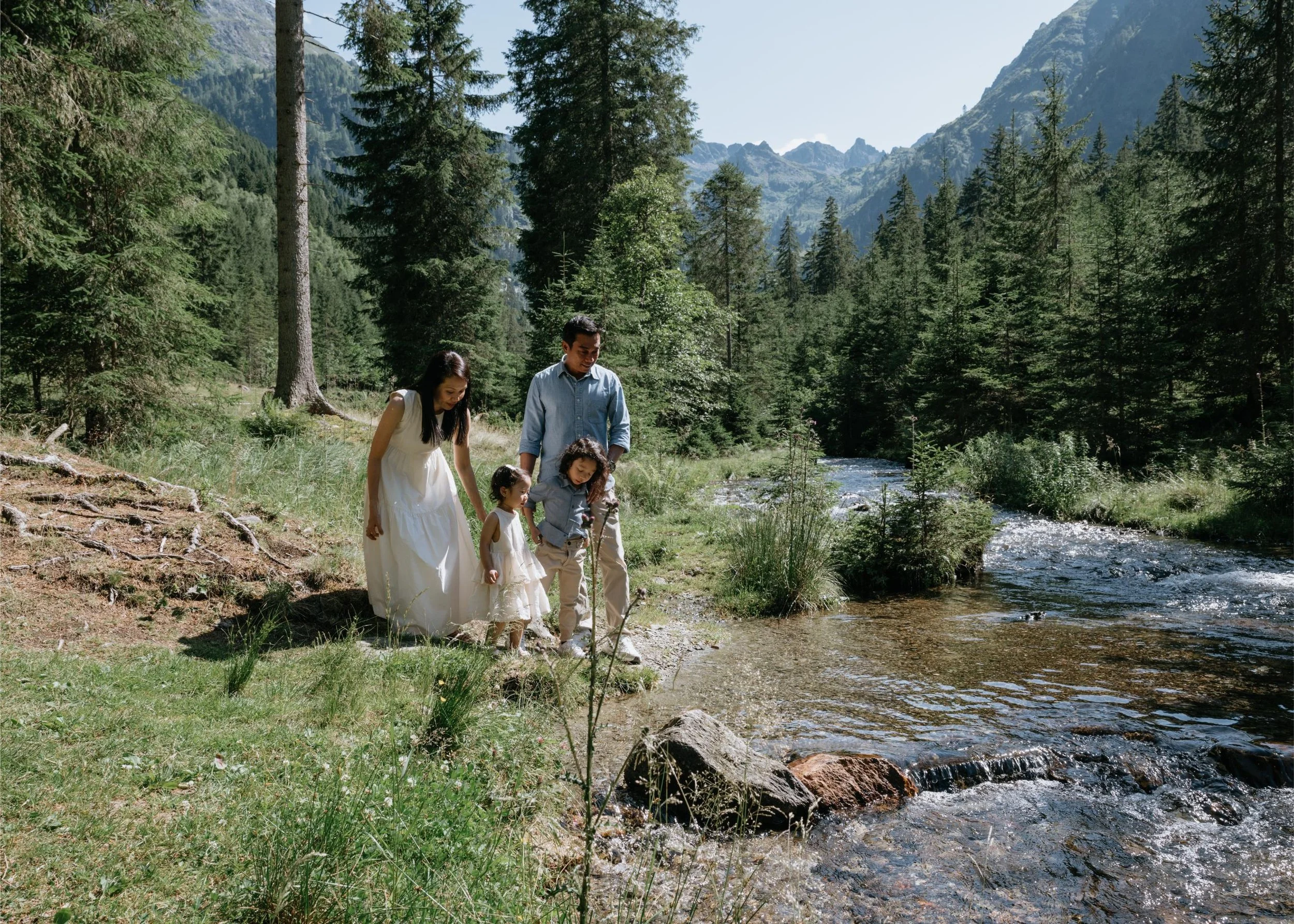 A family of four, consisting of a woman, a man, and two young girls, enjoying a walk along a river in a forested mountain landscape on a sunny day.