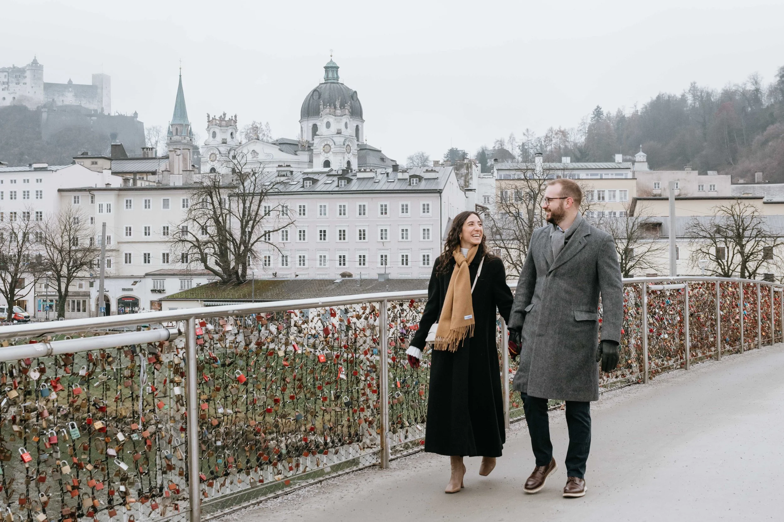 A smiling couple walking along a bridge with love locks in a European city, with historic buildings and a hilltop castle in the background.