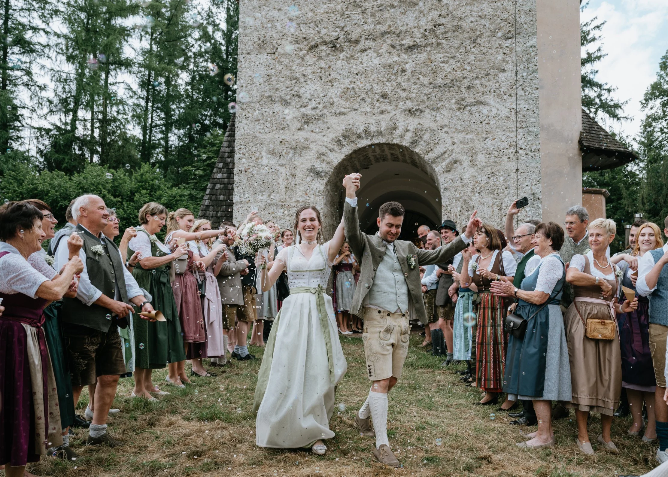A newlywed couple is celebrating as they walk through a crowd of friends and family outside an old stone building. The bride is wearing a traditional white dress holding a bouquet, and the groom is dressed in traditional Bavarian attire. The crowd is