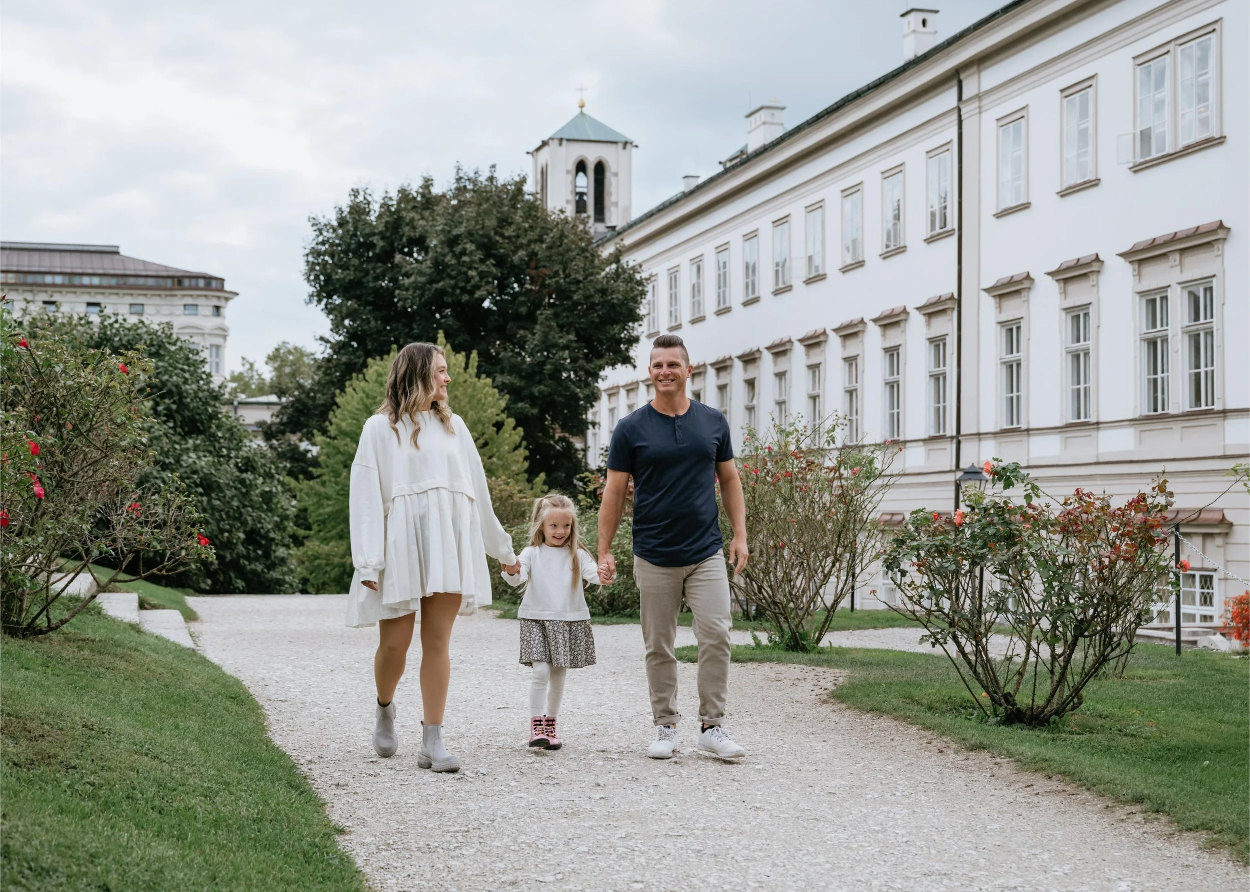 Family of four walking on a gravel path in a park, holding hands and smiling, with a white building and trees in the background.