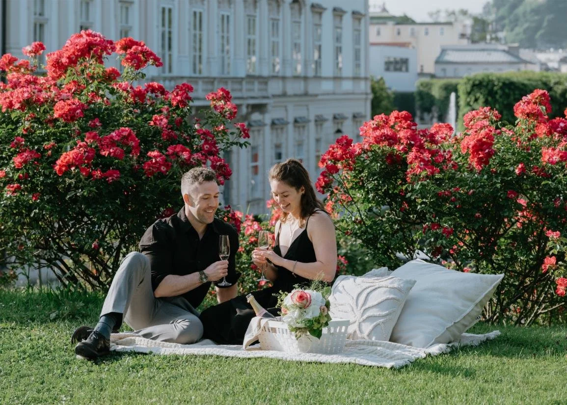 A man and woman sitting on a grassy area during a picnic, holding wine glasses, with pink flowering bushes and a cityscape in the background.