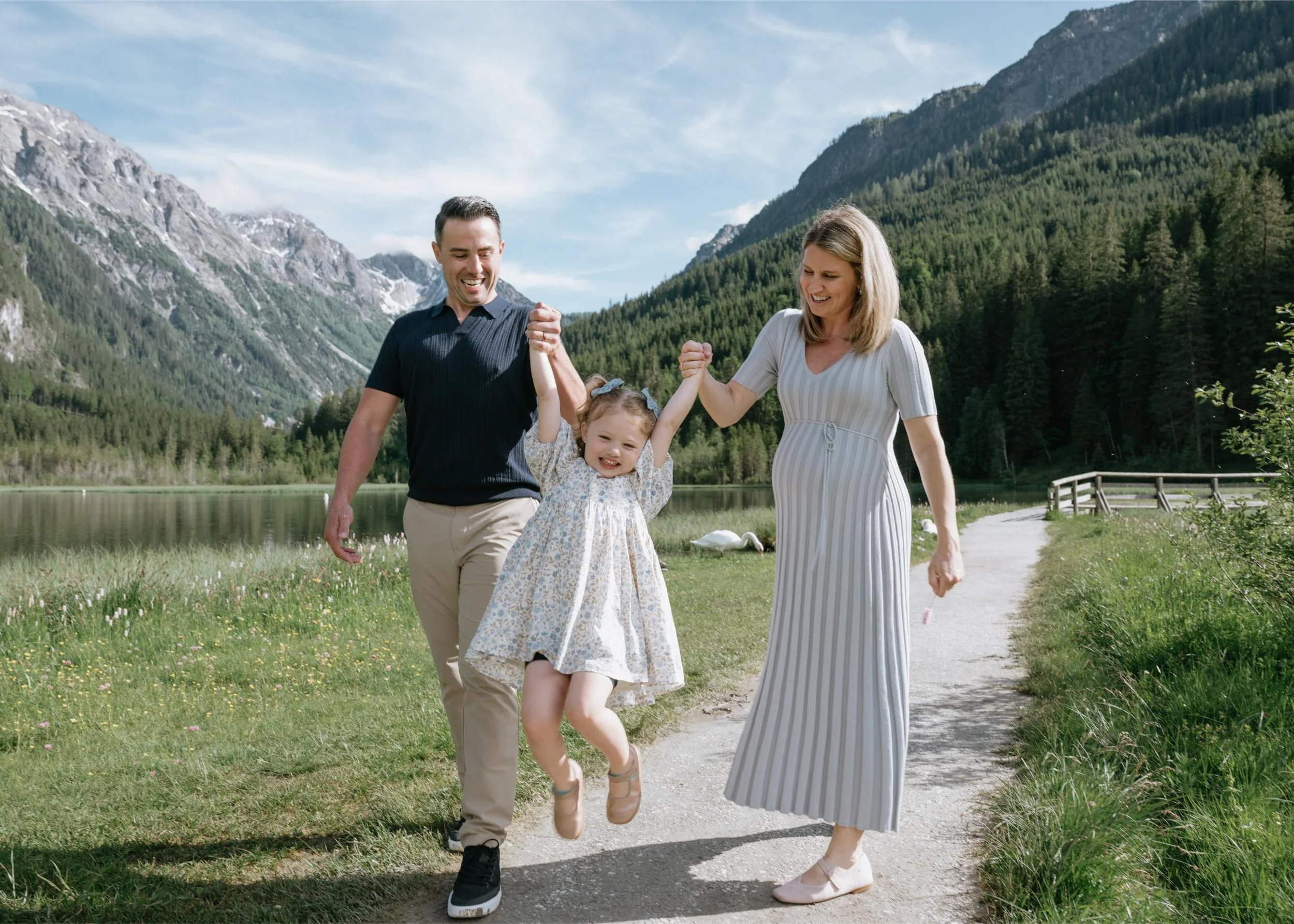 A happy family of three, a father, mother, and young daughter, enjoying a sunny day outdoors near a lake with mountains and trees in the background. The father and mother are holding the daughter's hands as she jumps in the air.
