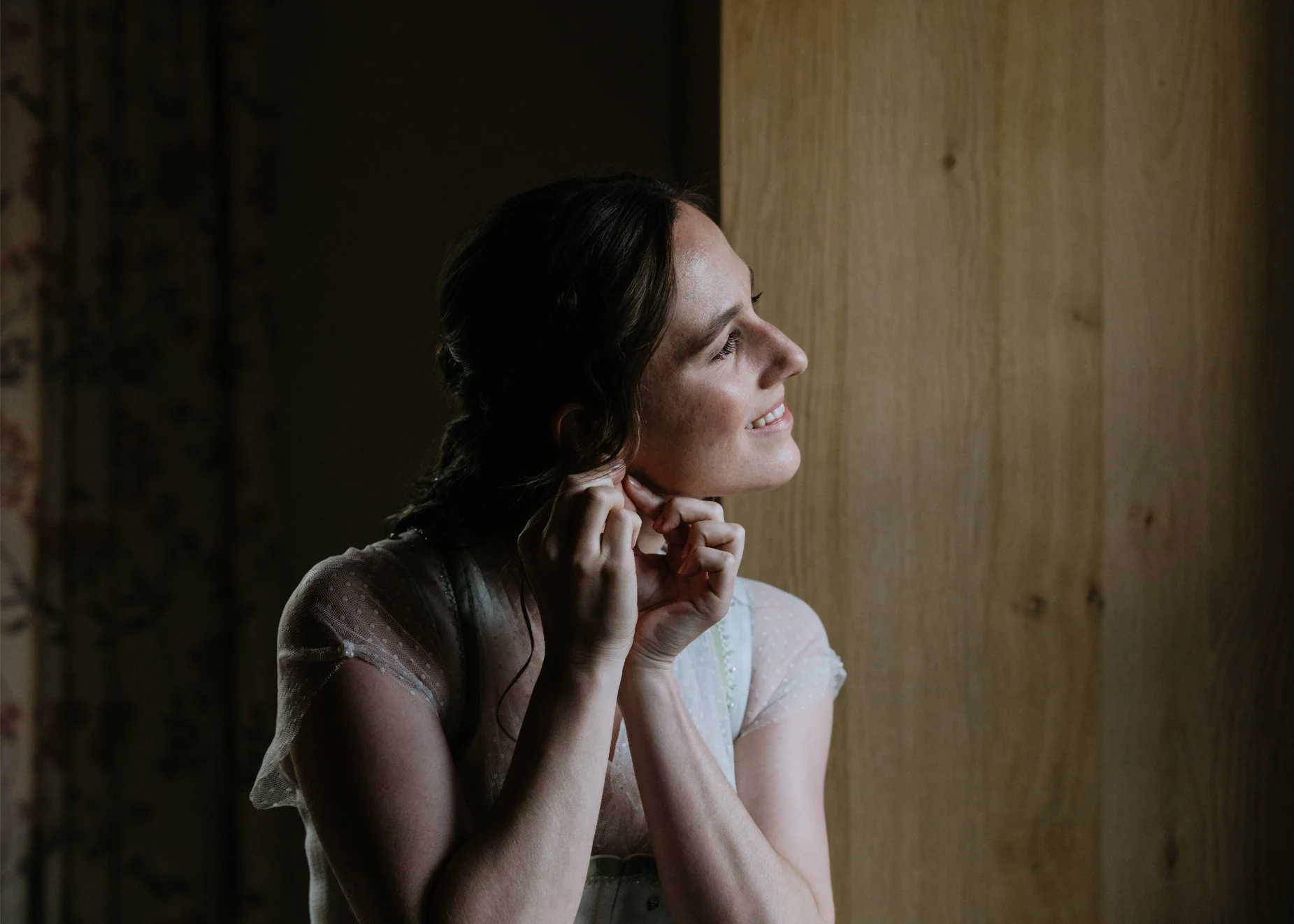 A woman with dark hair smiles and adjusts her earring as she looks out a window with natural light illuminating her face.