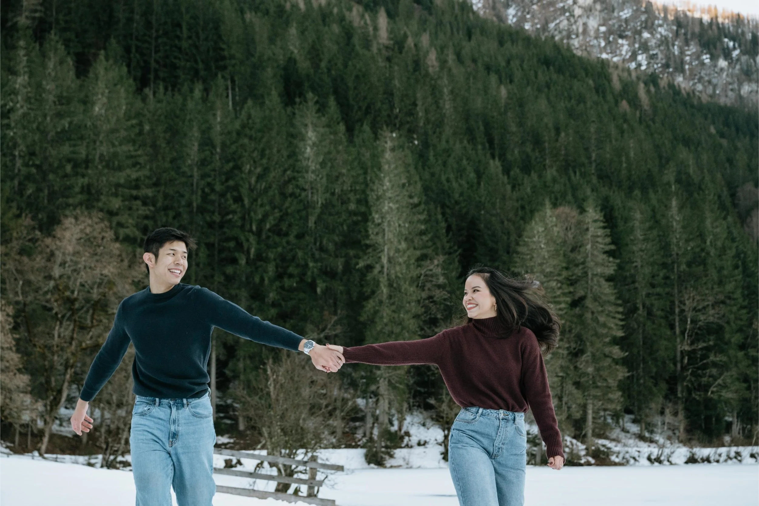 A young couple holding hands and smiling while walking in a snowy landscape with a dense green forest and mountain background.
