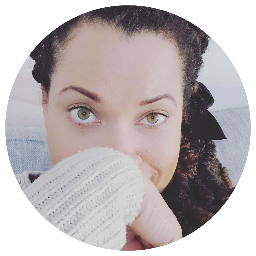 Close-up photo of a woman with curly hair, green eyes, wearing a white knitted sweater, partially covering her face with her hand.