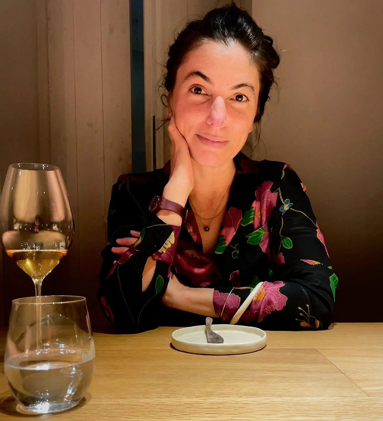 A woman with dark hair in a braid, wearing a black floral blouse, sitting at a wooden table with a glass of white wine, a glass of water, and an empty dish in front of her, smiling gently at the camera.