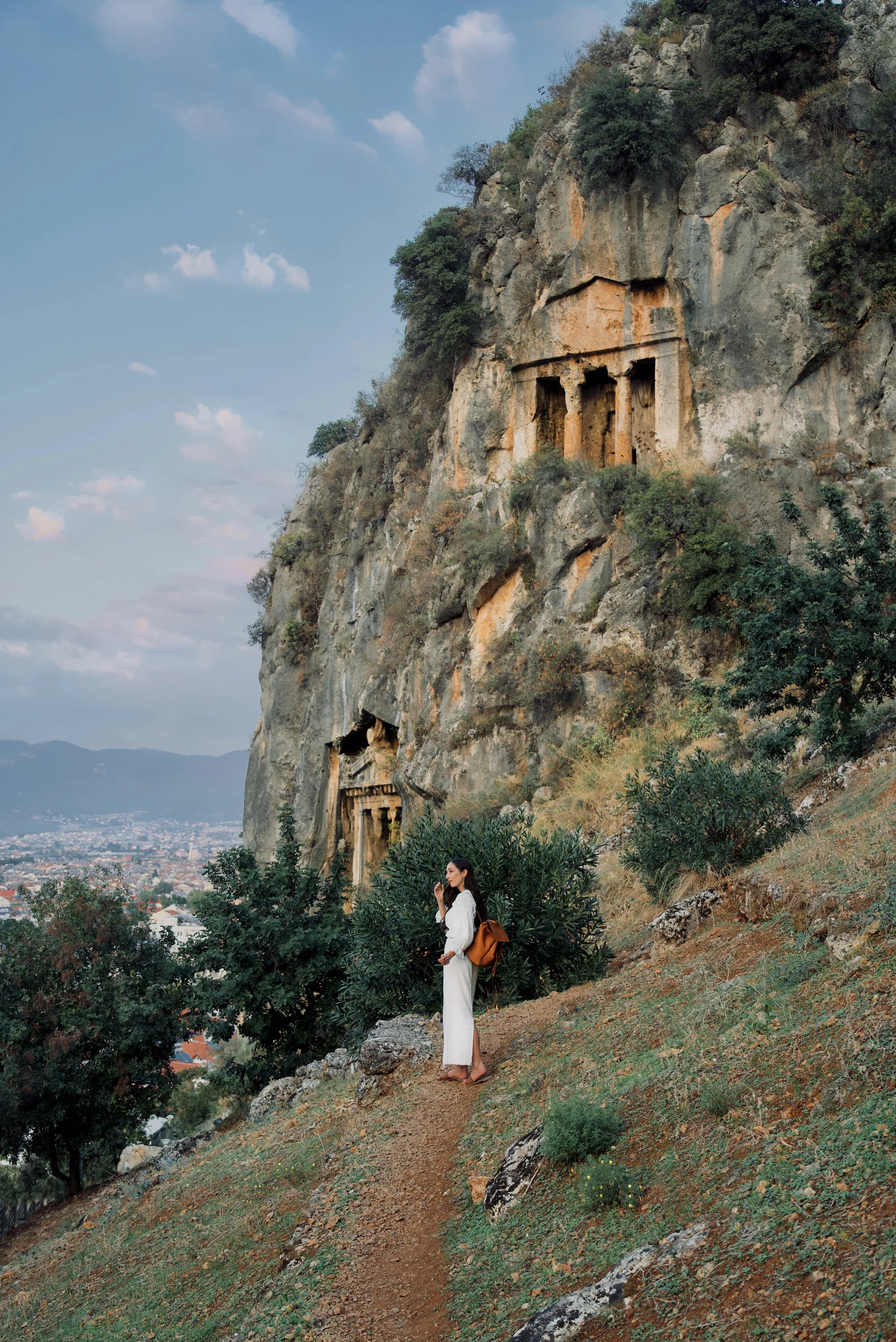 A girl in a white linen outfit stands on the hills with the Amyntas Rock Tombs in the background near Fethiye, Türkiye (Turkey)