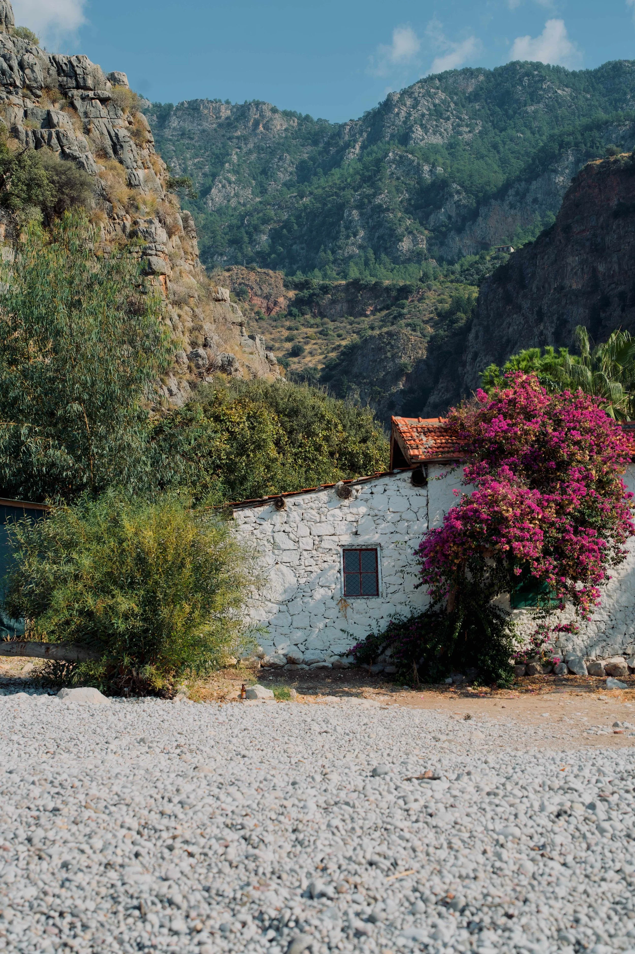 An aged white brick home near the beach of the Butterfly Valley, Türkiye (Turkey). Cliff tops and a luscious valley strecthes into the background behind the house.