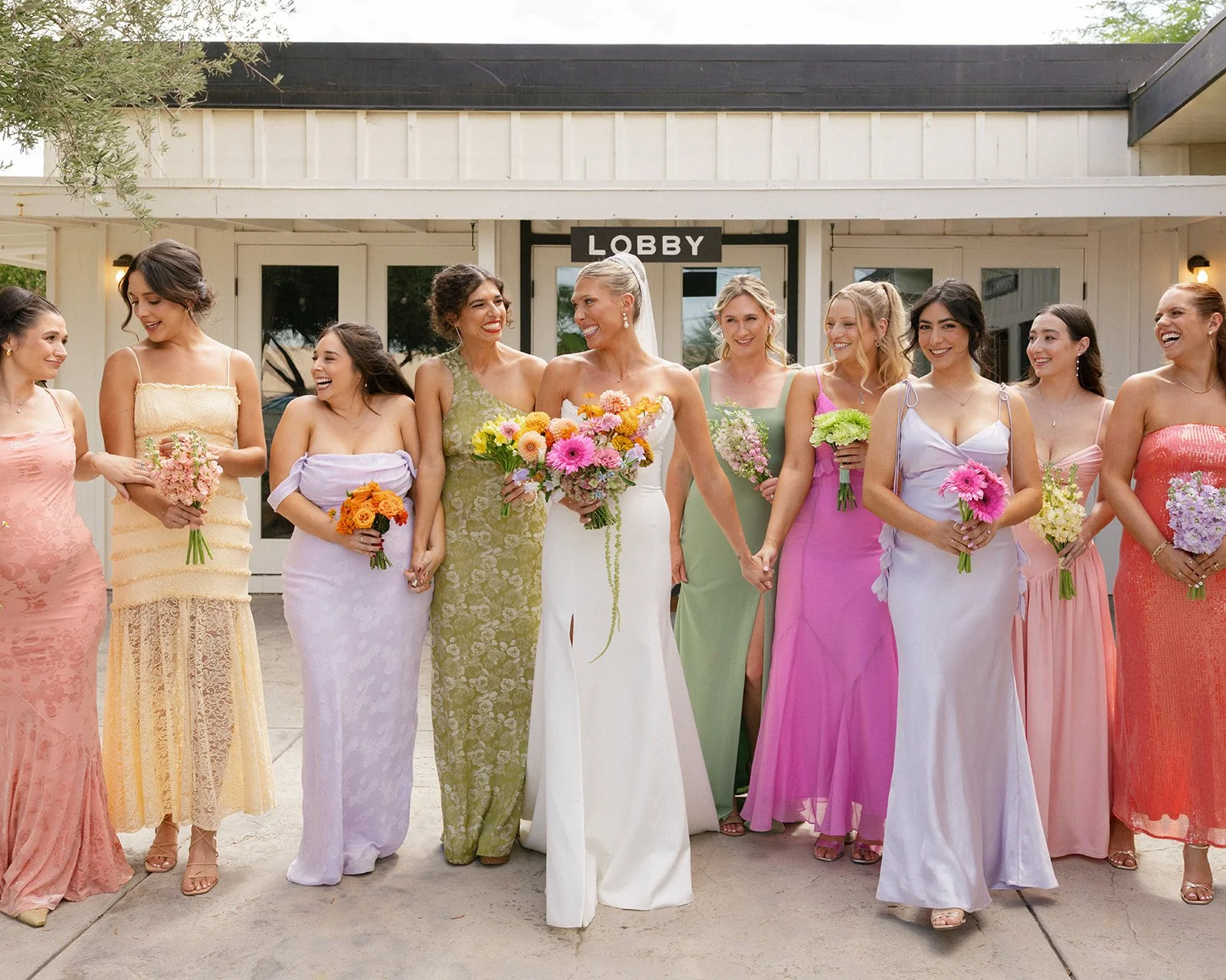 A bride dressed in white with her bridesmaids in colorful dresses all holding colorful bouquets.
