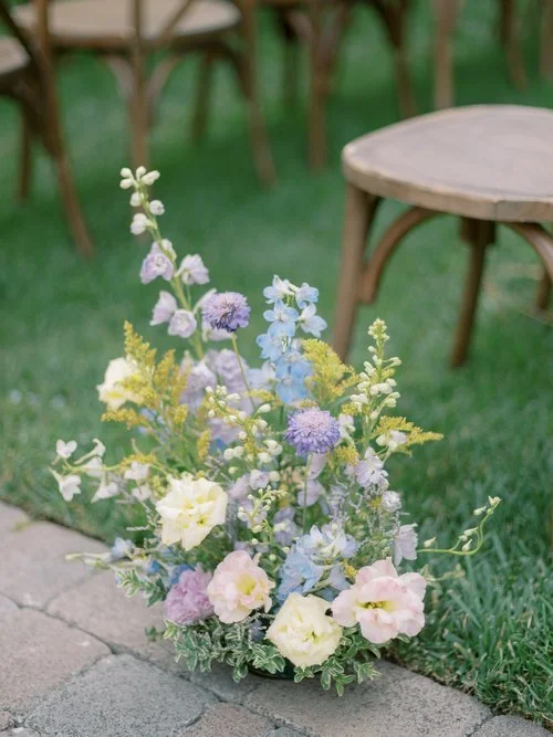 Pastel floral arrangement placed next to wooden chairs for outdoor wedding.