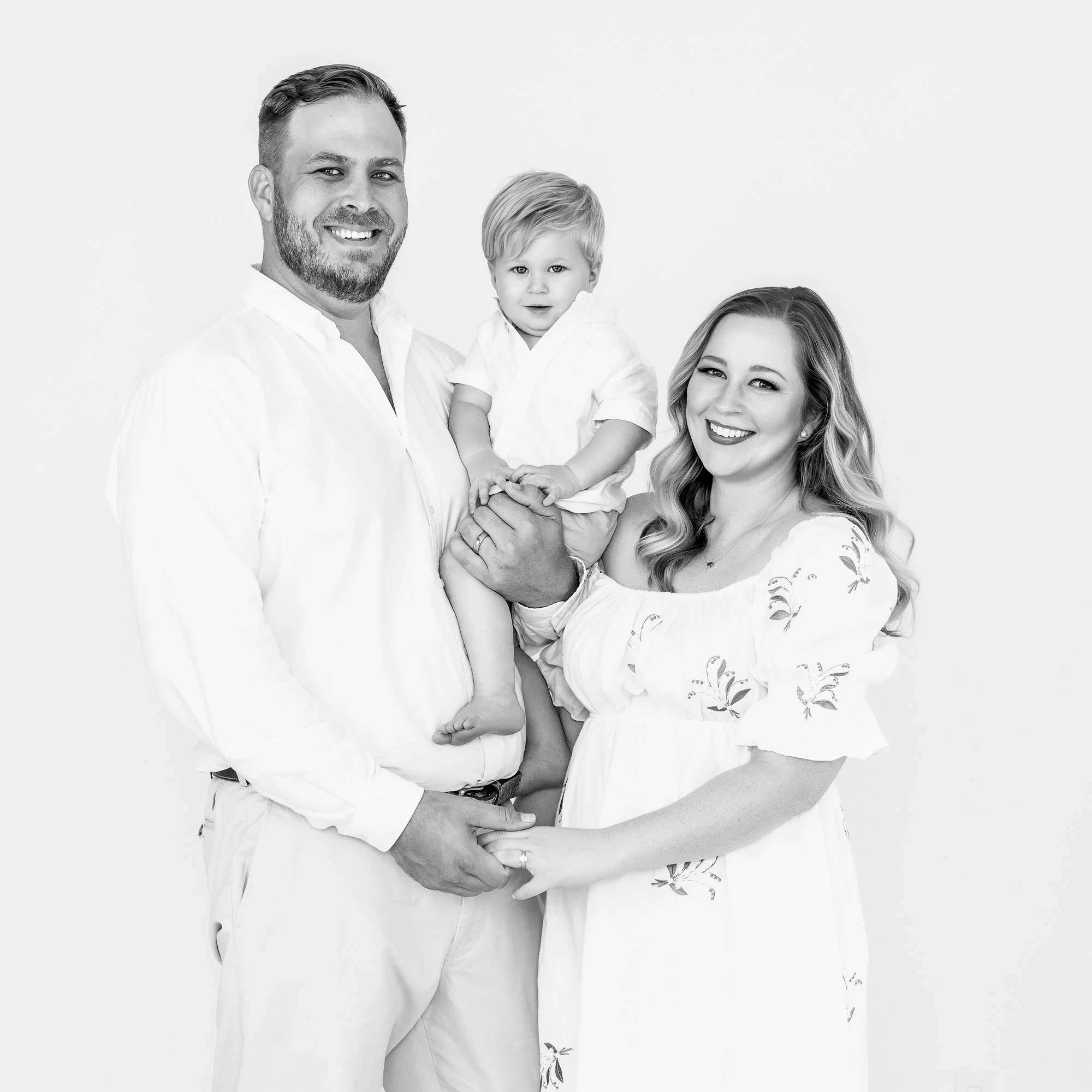 Black and white studio portrait of an expecting mother, father and toddler son dressed in white, McAllen TX maternity and family photographer