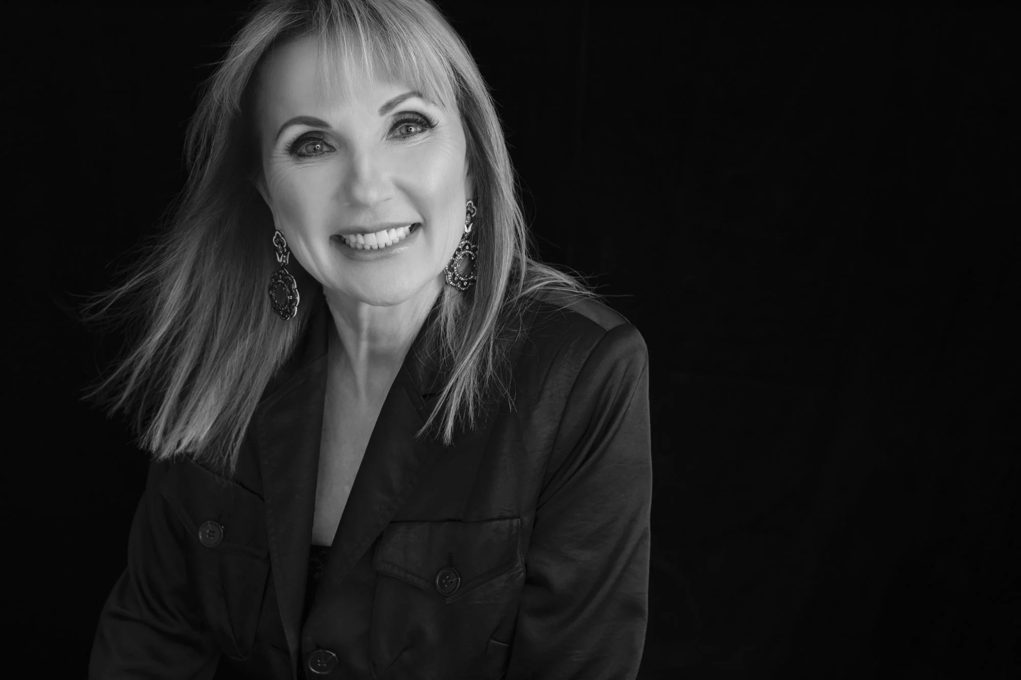 Black and white studio portrait of a smiling woman with long hair and statement earrings against a black backdrop, McAllen TX women's portrait photographer