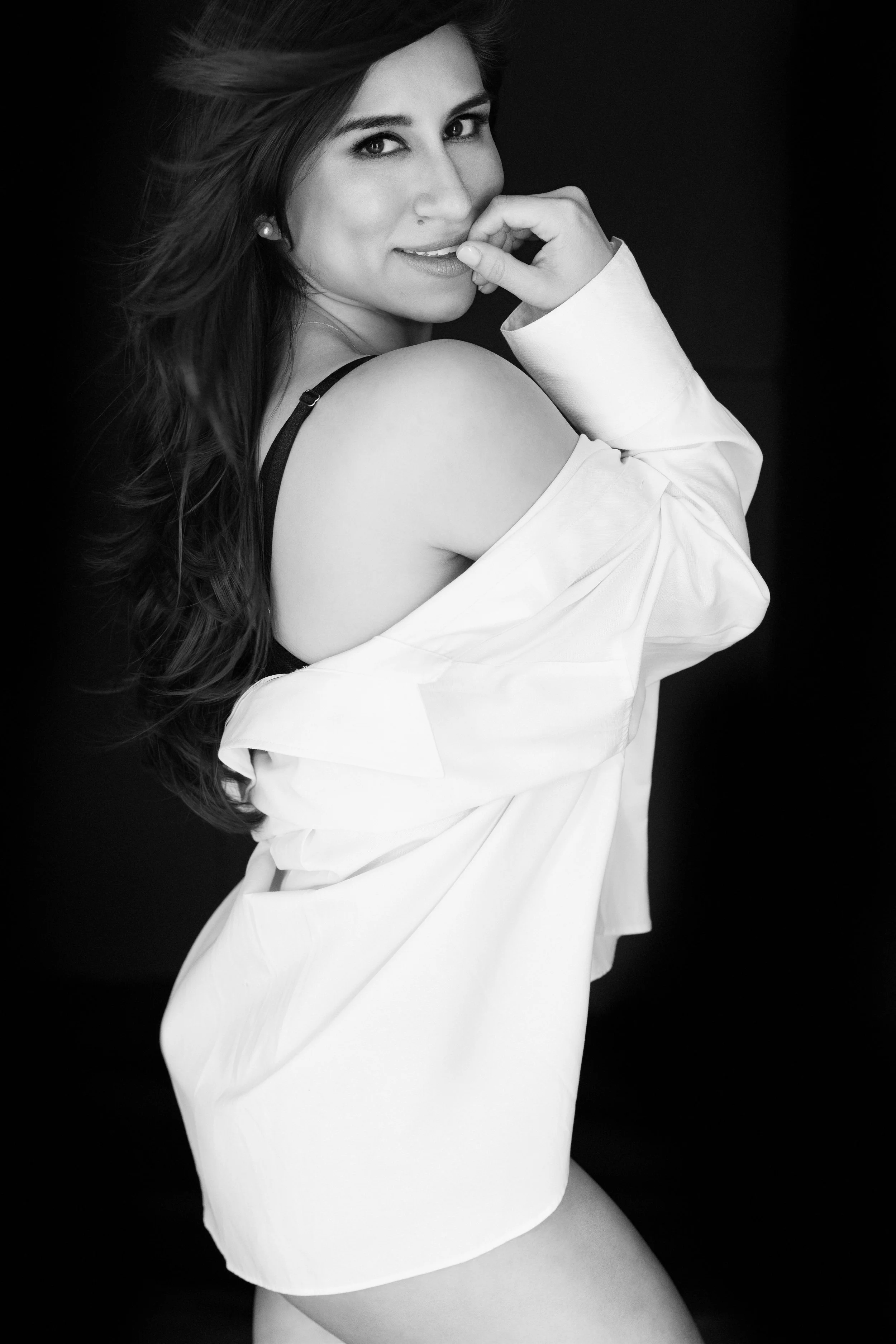 Black and white image of Nicole Gates Photography's Unbuttoned Photography campaign. The woman is wearing sexy black lingerie and a large white shirt. she is confident and coy. looking at the camera and playfully covering her mouth with her hand.