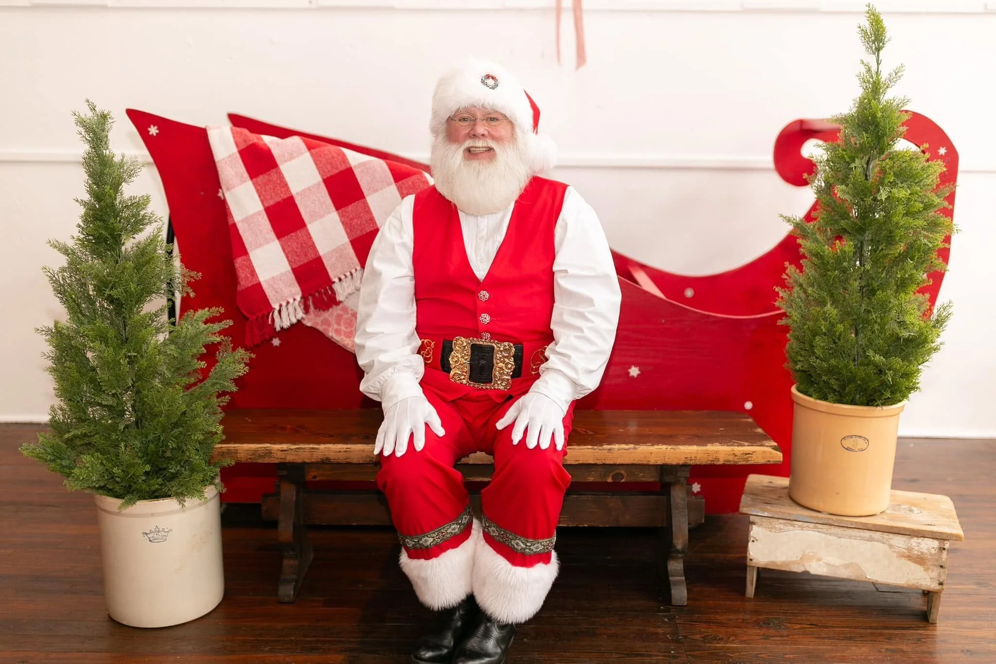 Santa sitting on a wooden bench with a red sleigh behind him. There are two small pine trees on either side of the bench.