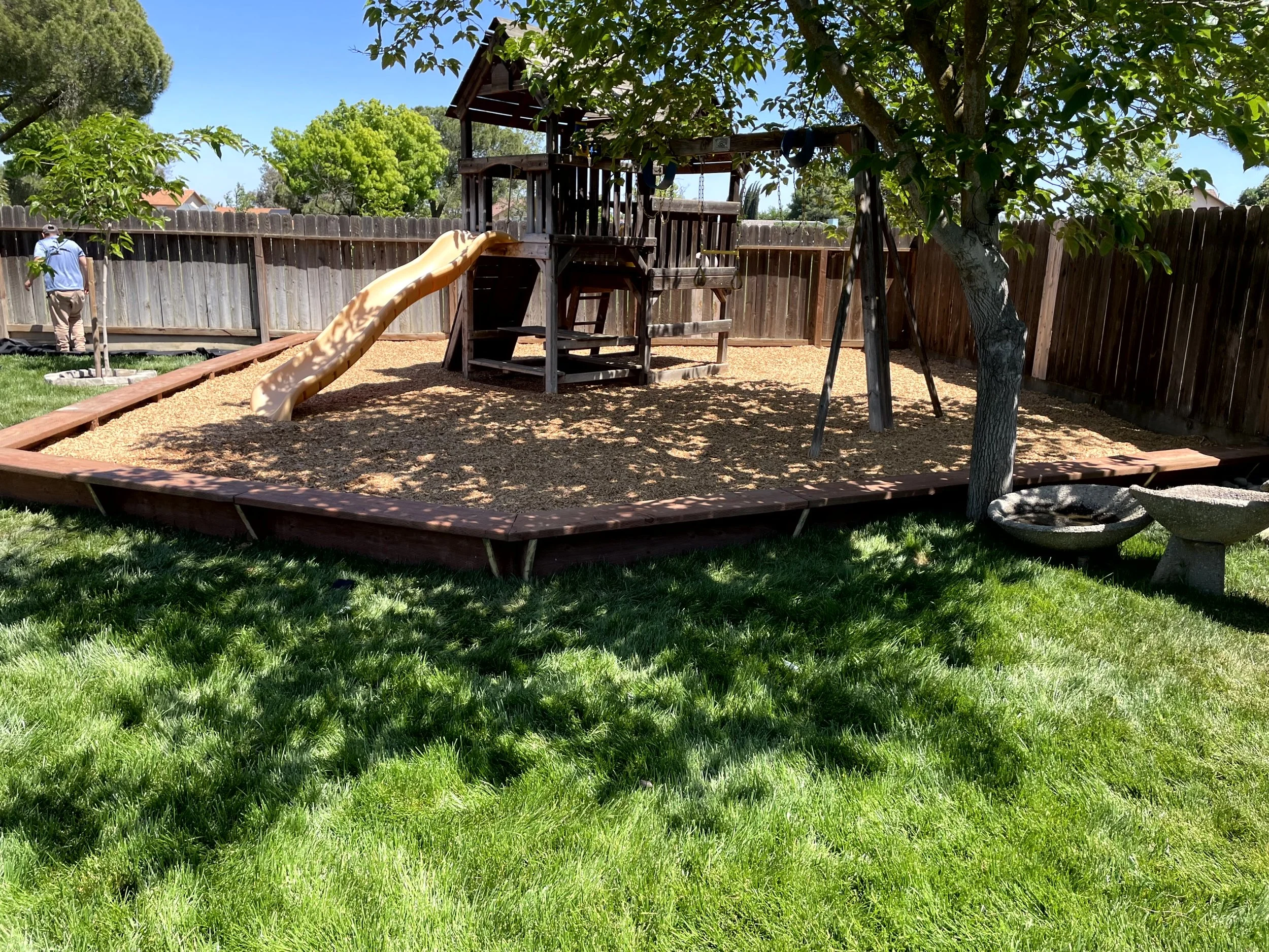 A backyard playground with a wooden play structure, slide, swings, and sandpit, surrounded by a wooden fence and green grass, with trees providing shade.