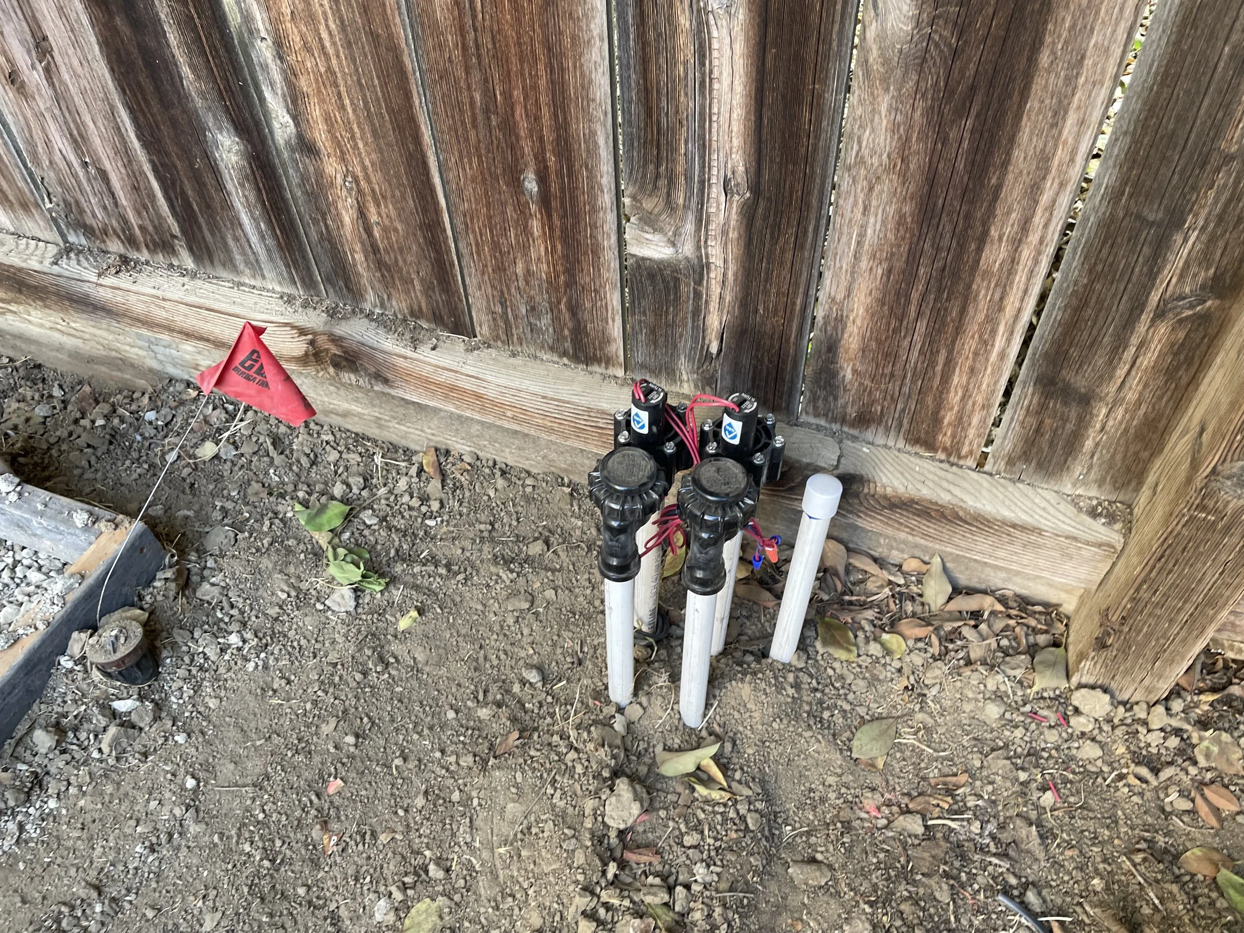 underground sprinkler system manifold with three black zone valves and white PVC pipes next to a wooden fence, on dirt ground with scattered leaves and a small red flag nearby.