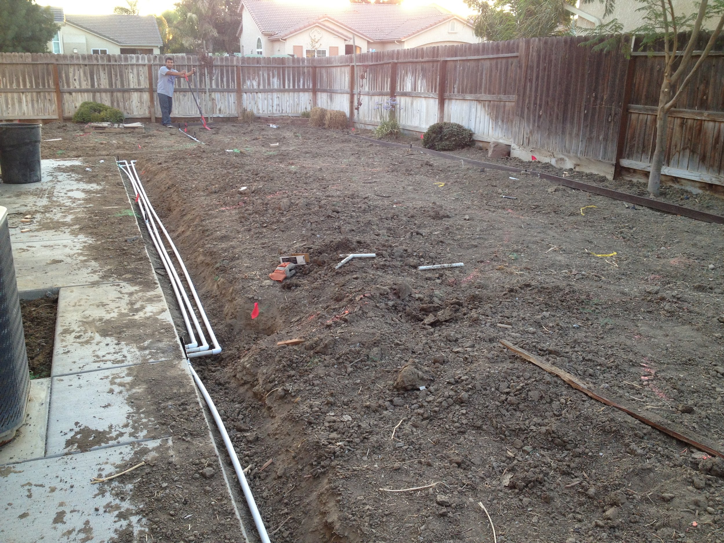 A backyard with dirt being leveled and prepared for construction, with a man using a tool, fences, and some trees and houses in the background.